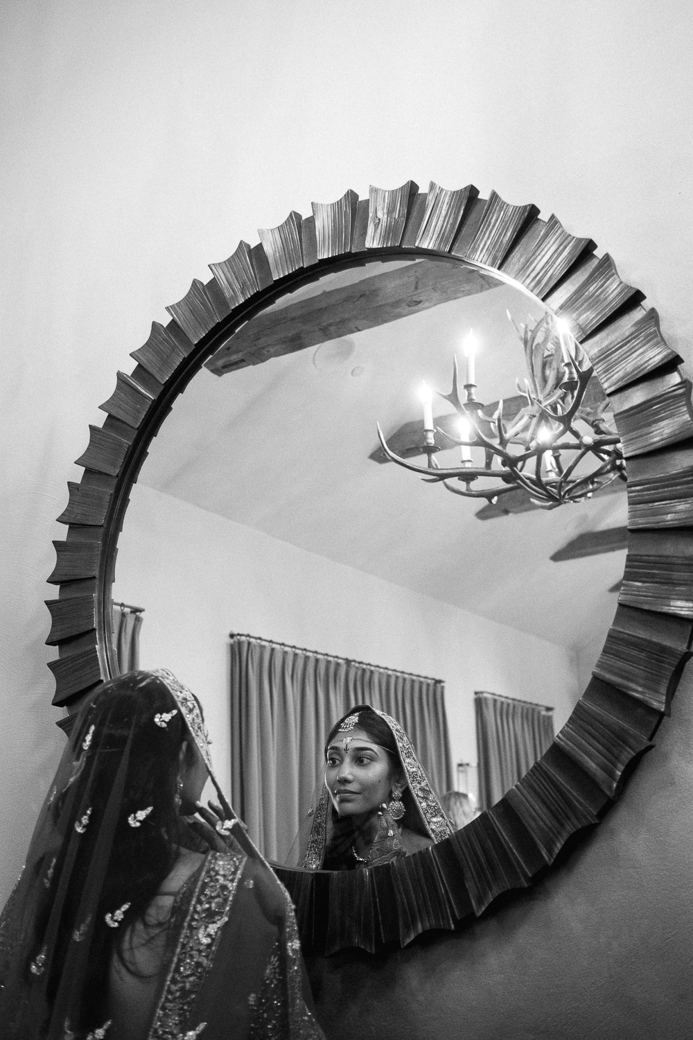 Woman dressed in traditional Indian attire looking at her reflection in a decorative wall mirror with a chandelier visible above.
