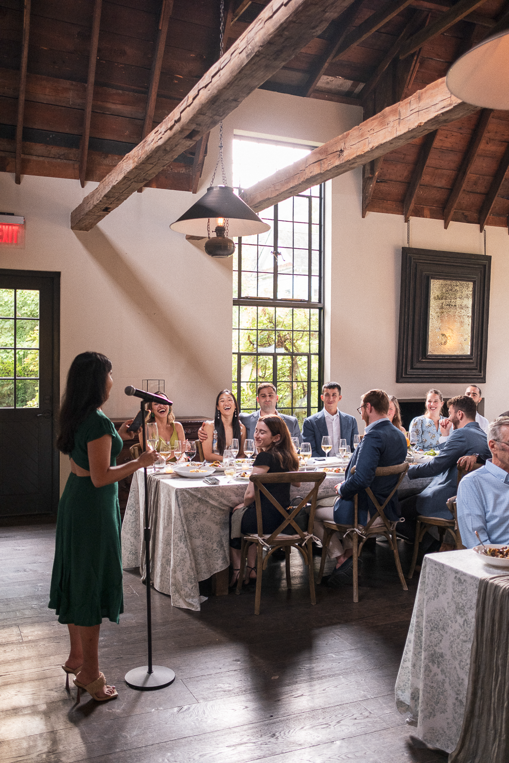 A woman in a green dress giving a speech or toast to a group of people sitting at a round banquet table in a rustic dining room with wooden beams and large windows.