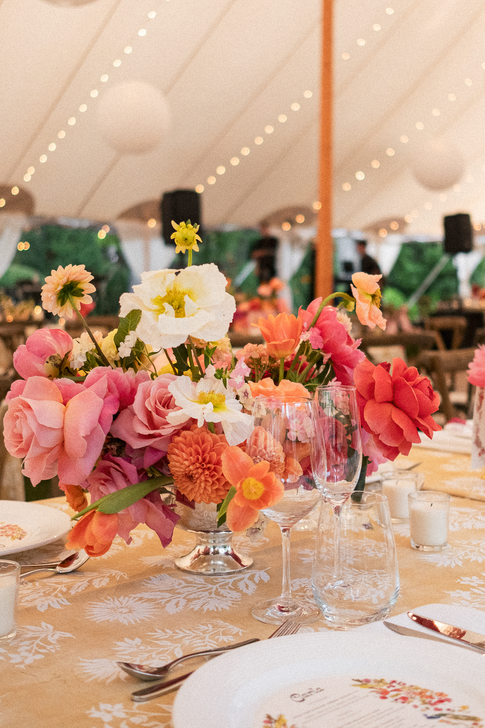 A floral centerpiece with pink, white, and orange flowers on a decorated table at a wedding or event under a tent.