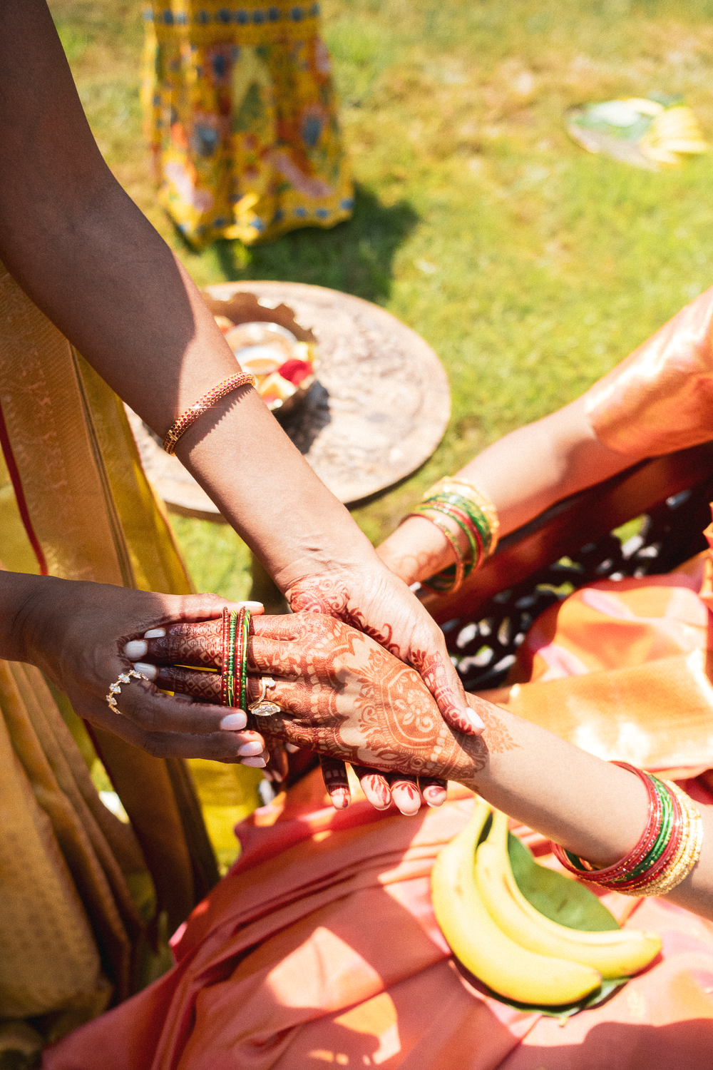 Close-up of two women holding hands during an outdoor traditional Indian ceremony, with henna on their hands and colorful bangles, a banana on their lap, and a decorated table in the background.