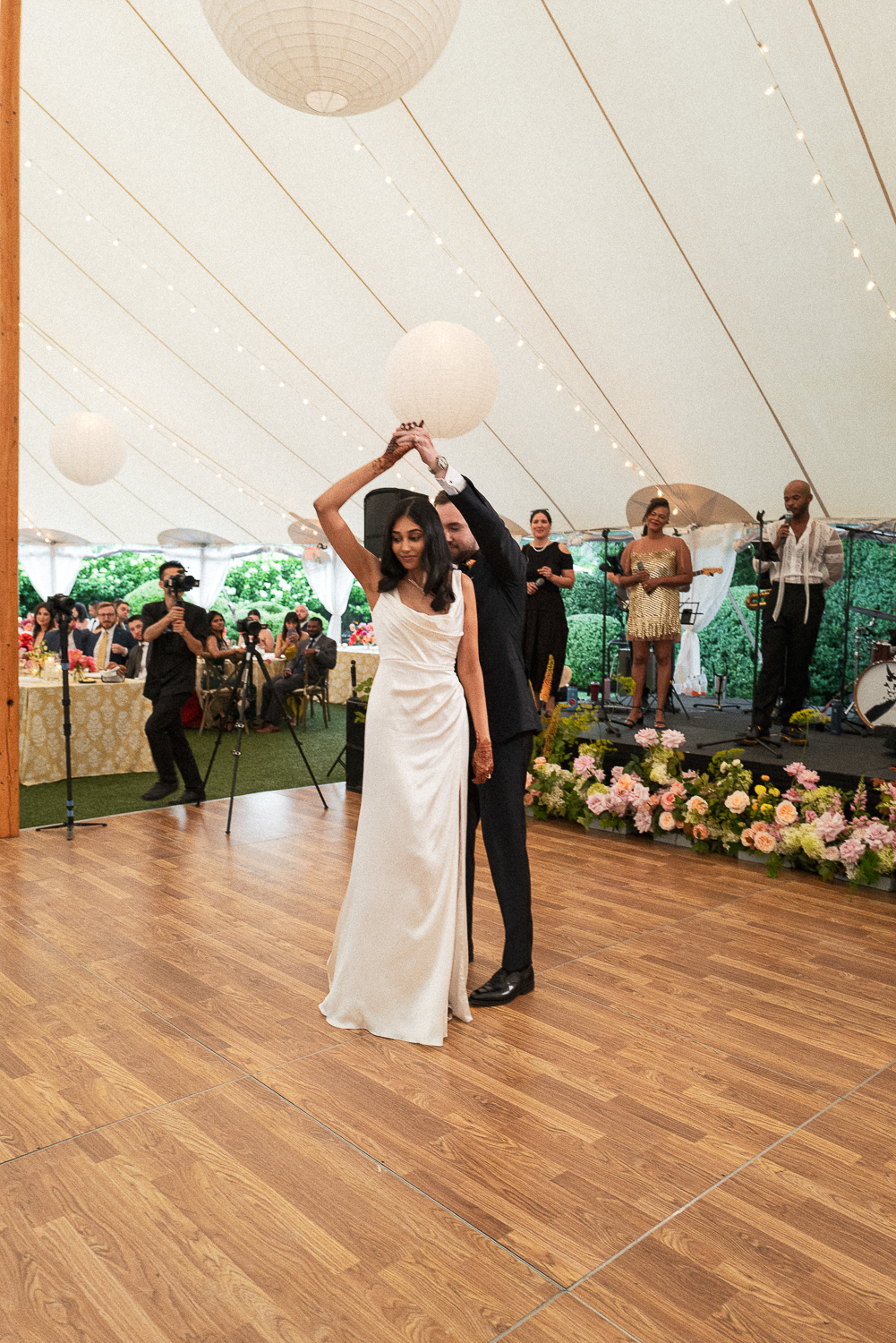 Couple dancing at a wedding reception under a tent decorated with string lights and paper lanterns, with guests and a live band in the background.