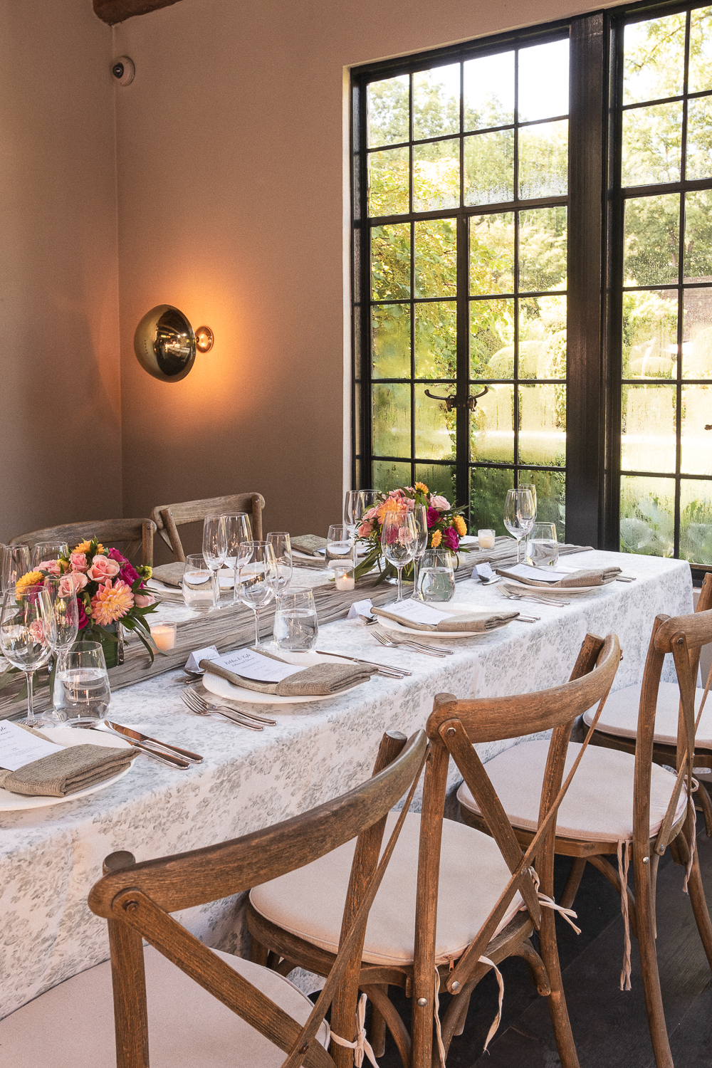 A decorated dining table set for a meal with pink, yellow, and purple flowers, surrounded by wooden chairs, near a large window with black framing, in a room with beige walls and a modern wall sconce.