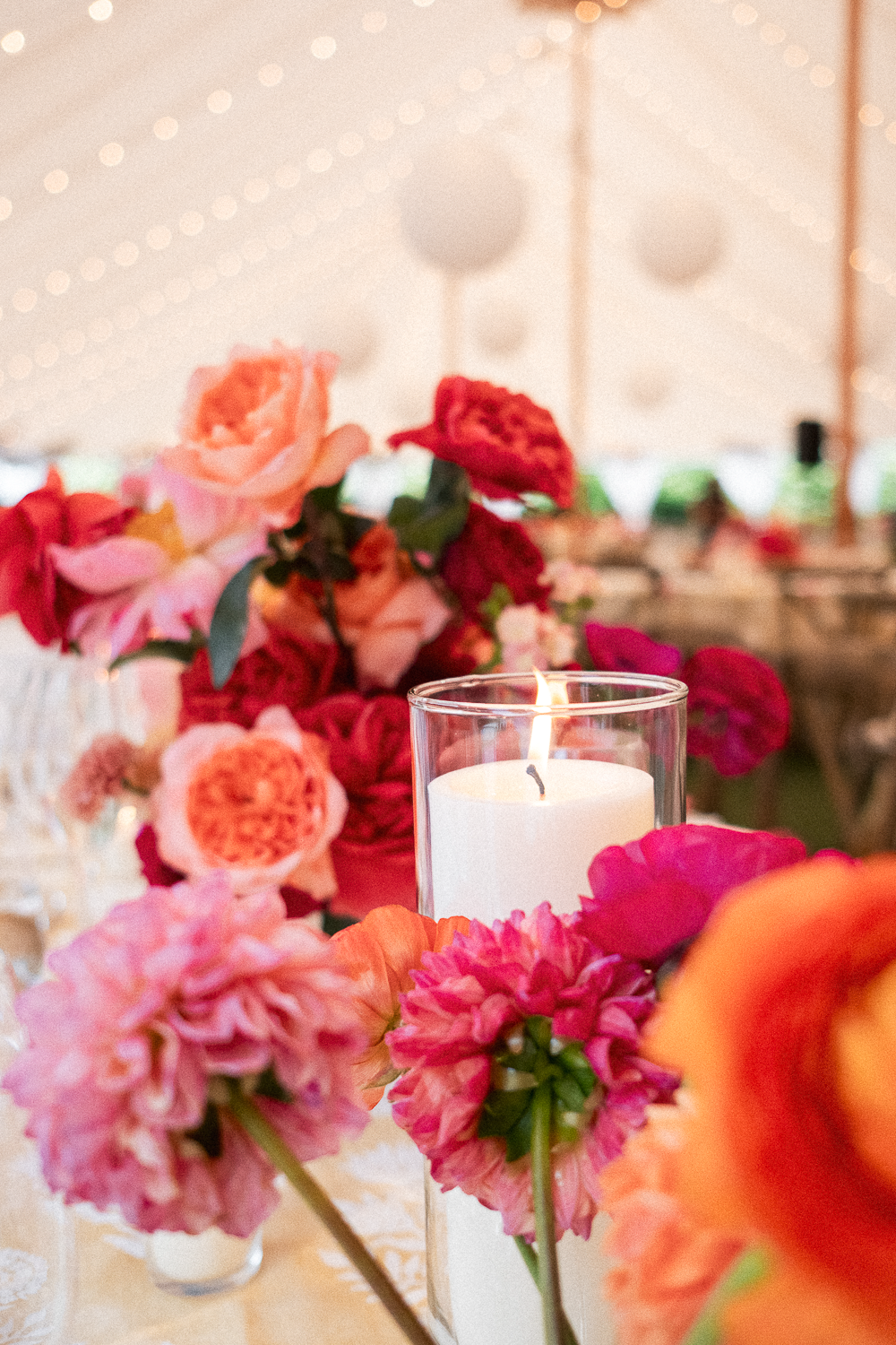 A floral centerpiece with pink, red, and orange flowers surrounding a lit white candle in a glass holder on a table at a decorated event space.