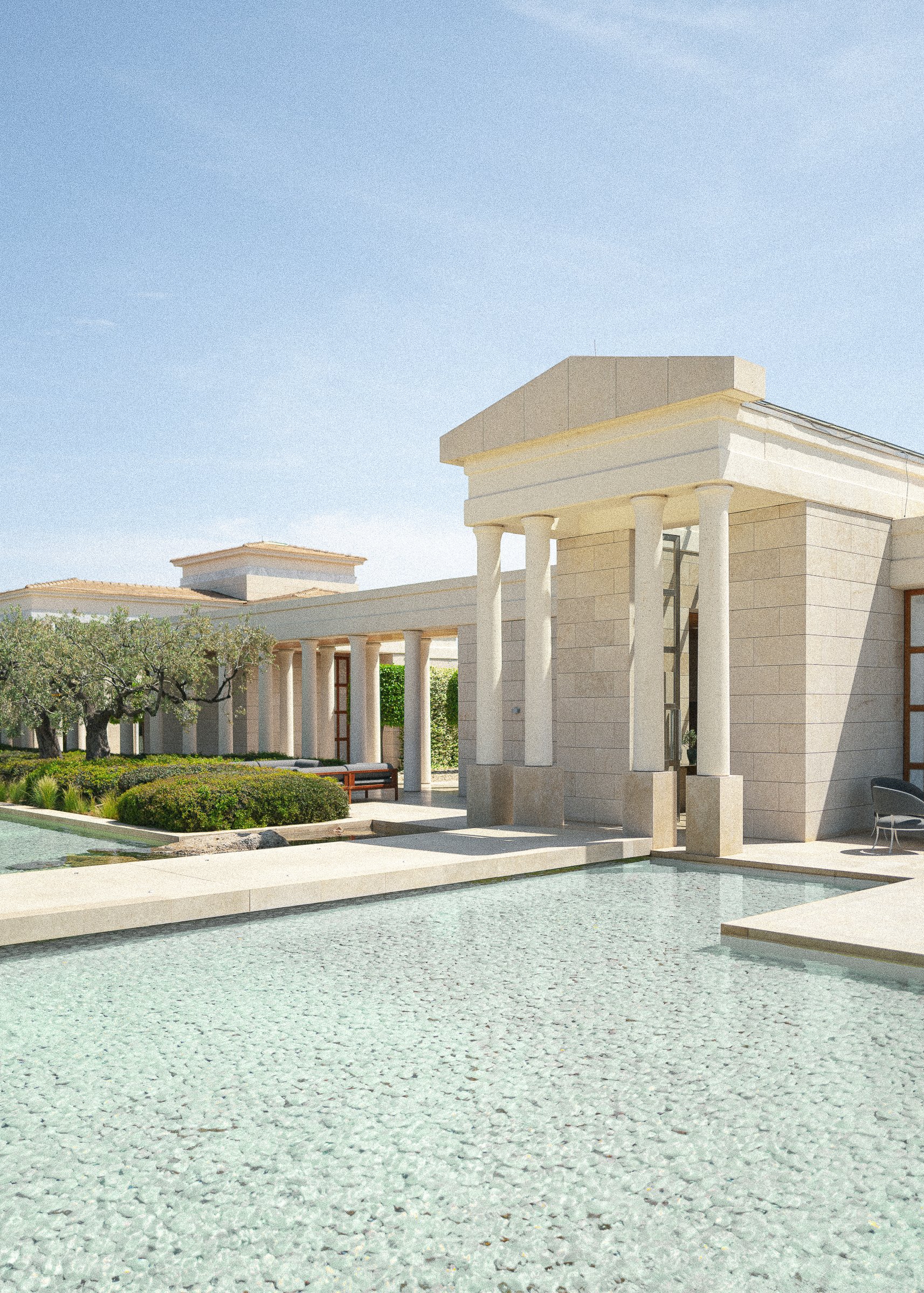 A modern building with beige stone walls and classical columns, surrounded by a shallow pool of water and greenery, under a clear blue sky.