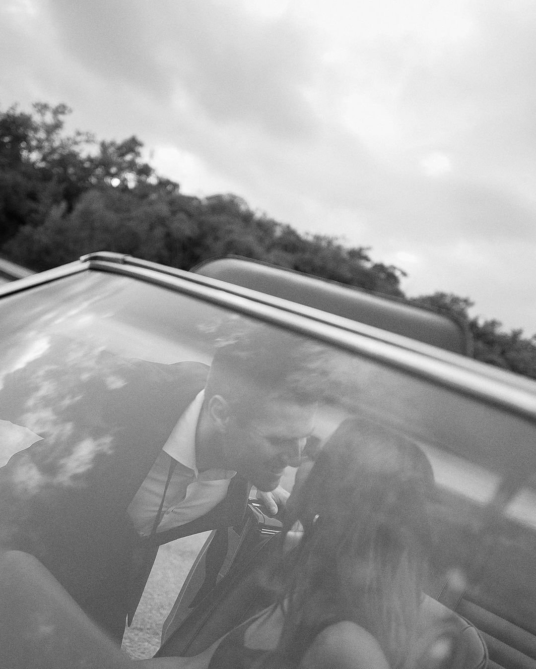 A black-and-white photo of a man looking at a woman through the window of a convertible car. The man has short hair and a beard, and the woman has long hair.