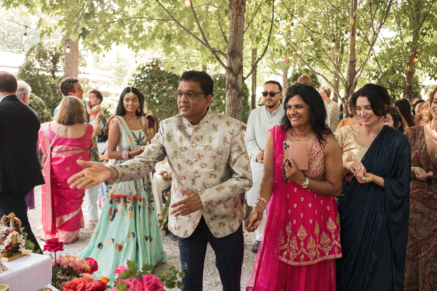 People at an outdoor celebration under a tree with string lights, including a man in a traditional cream-colored suit and women in colorful sarees, standing near a table with flowers and decorations.