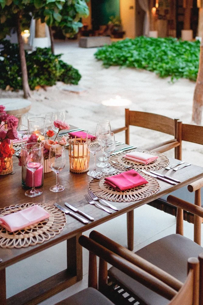 Set dining table with pink napkins, glassware, candles, and floral decorations in an outdoor veranda with greenery.
