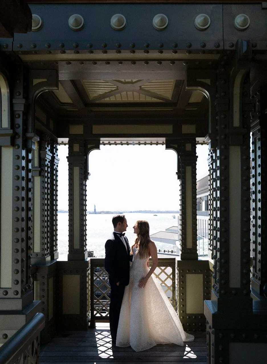 A bride and groom standing on a balcony overlooking the water in Cipriani South Street, framed by an ornate metal structure, with the Statue of Liberty visible in the background.
