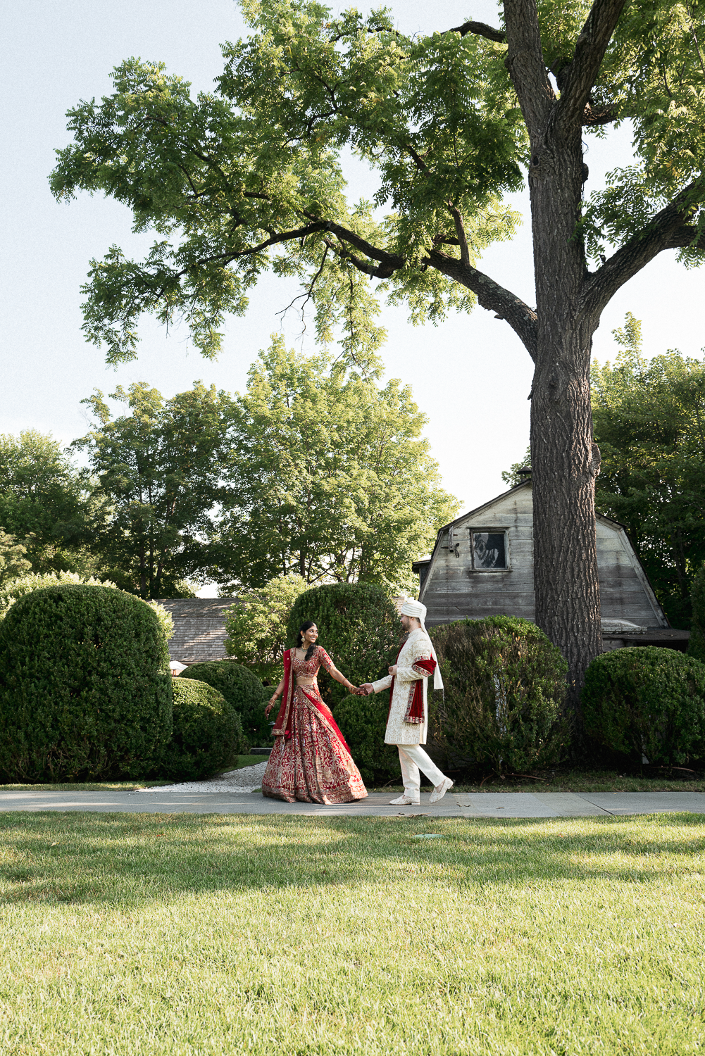 A couple dressed in traditional Indian wedding attire holding hands and walking on a sidewalk in a garden with trees and bushes.
