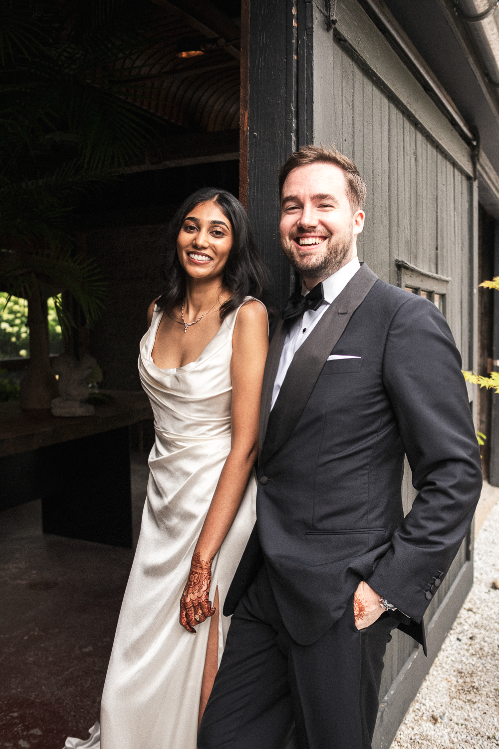 A happy couple dressed in formal attire, standing next to a black wooden wall, smiling at the camera.