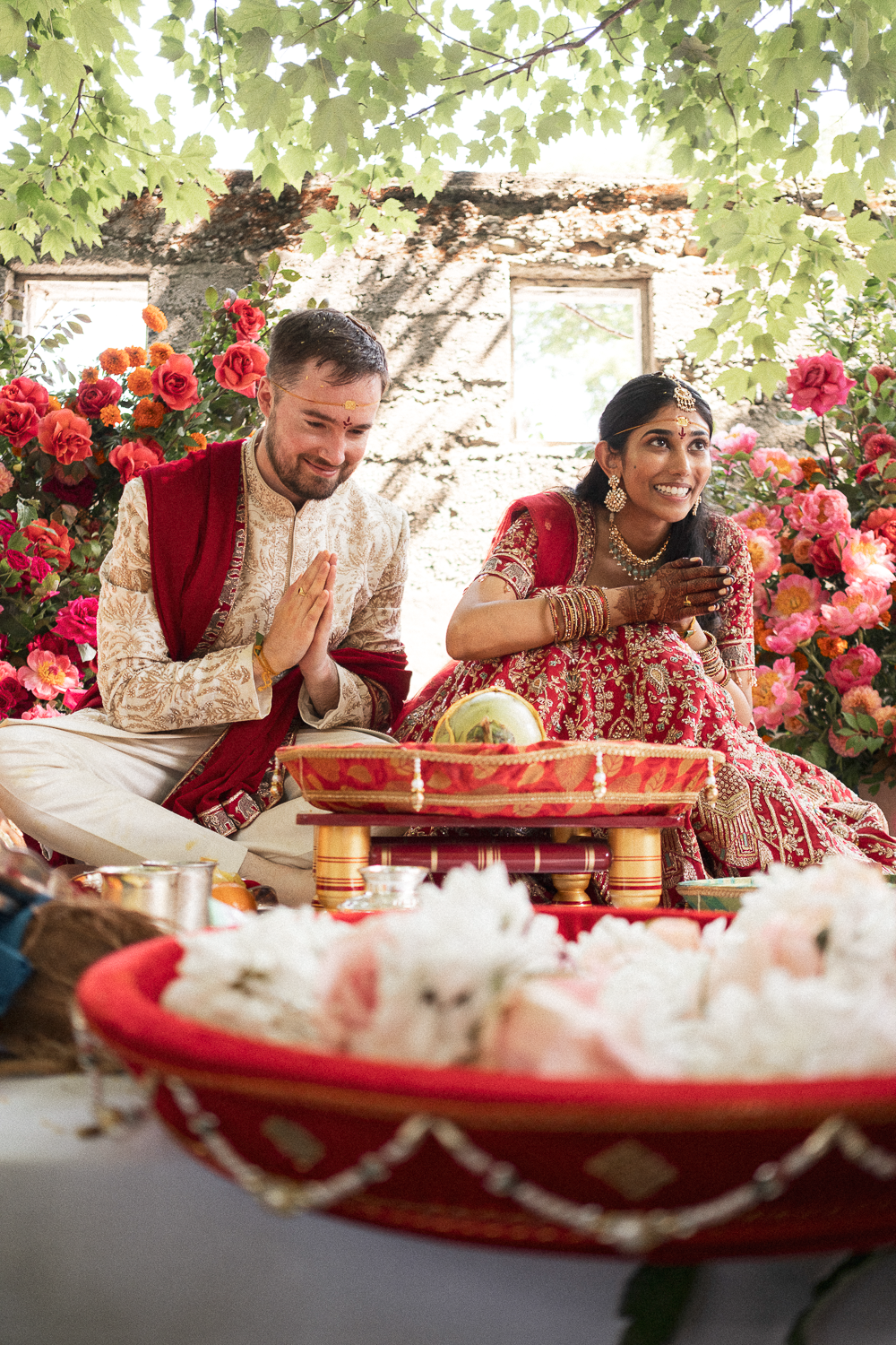A South Asian bride and a Caucasian groom sitting on the ground during a traditional Indian wedding ceremony, surrounded by pink and orange flowers, with the bride in a red and gold outfit and the groom in a beige and red outfit, offering prayers.
