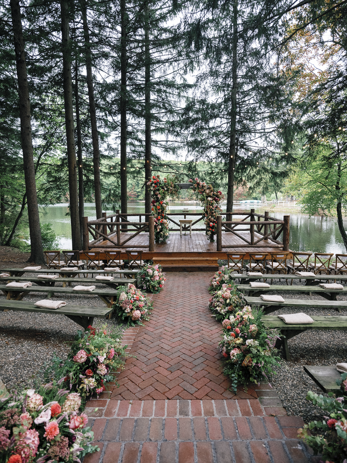 Outdoor wedding ceremony setup on a wooden platform with floral arches and seating in a wooded area with a lake in the background.