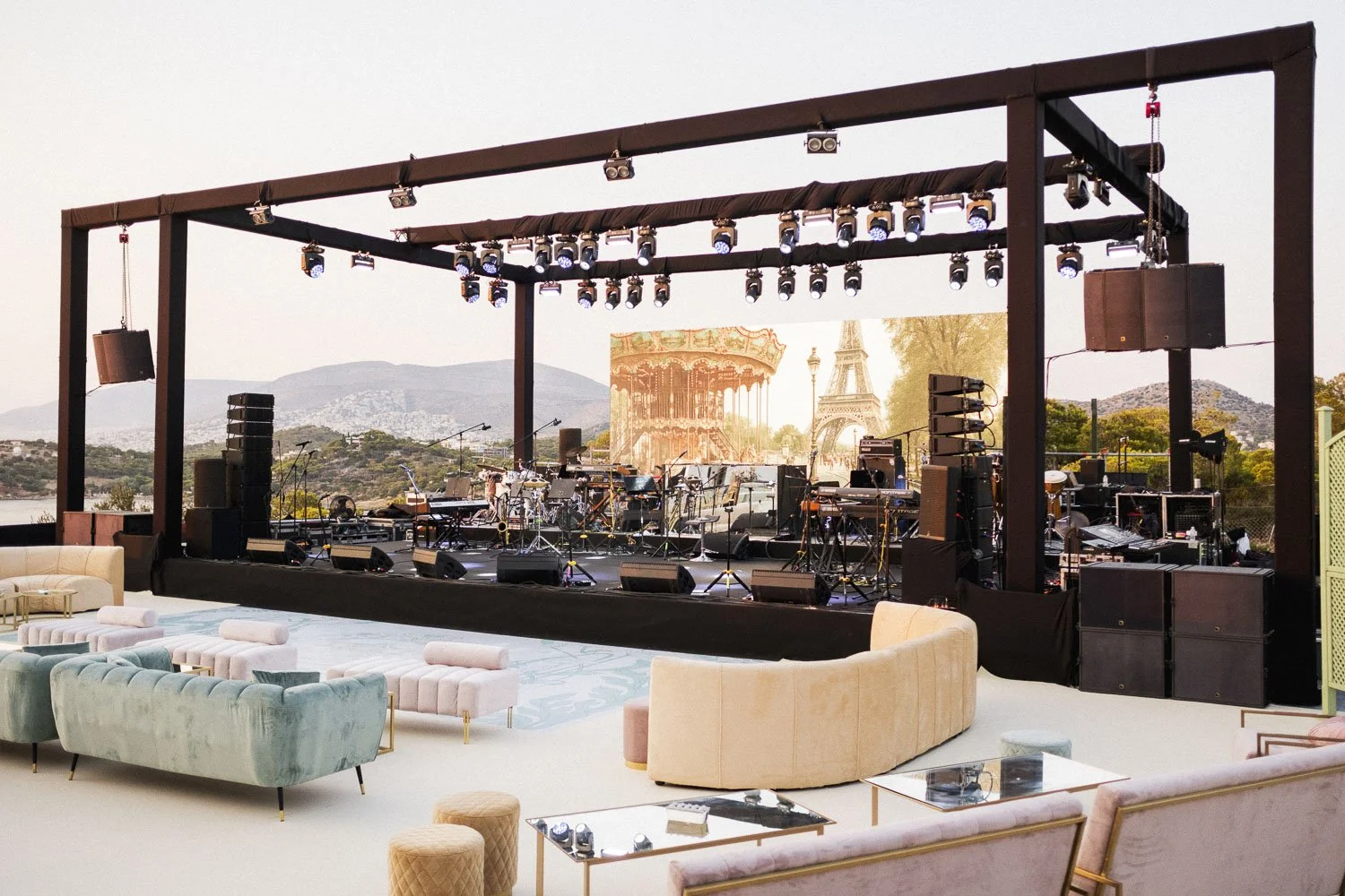 Outdoor stage with musical instruments and equipment set up, with Paris landmarks like the Eiffel Tower and carousel in the background, and plush chairs and sofas in the foreground.
