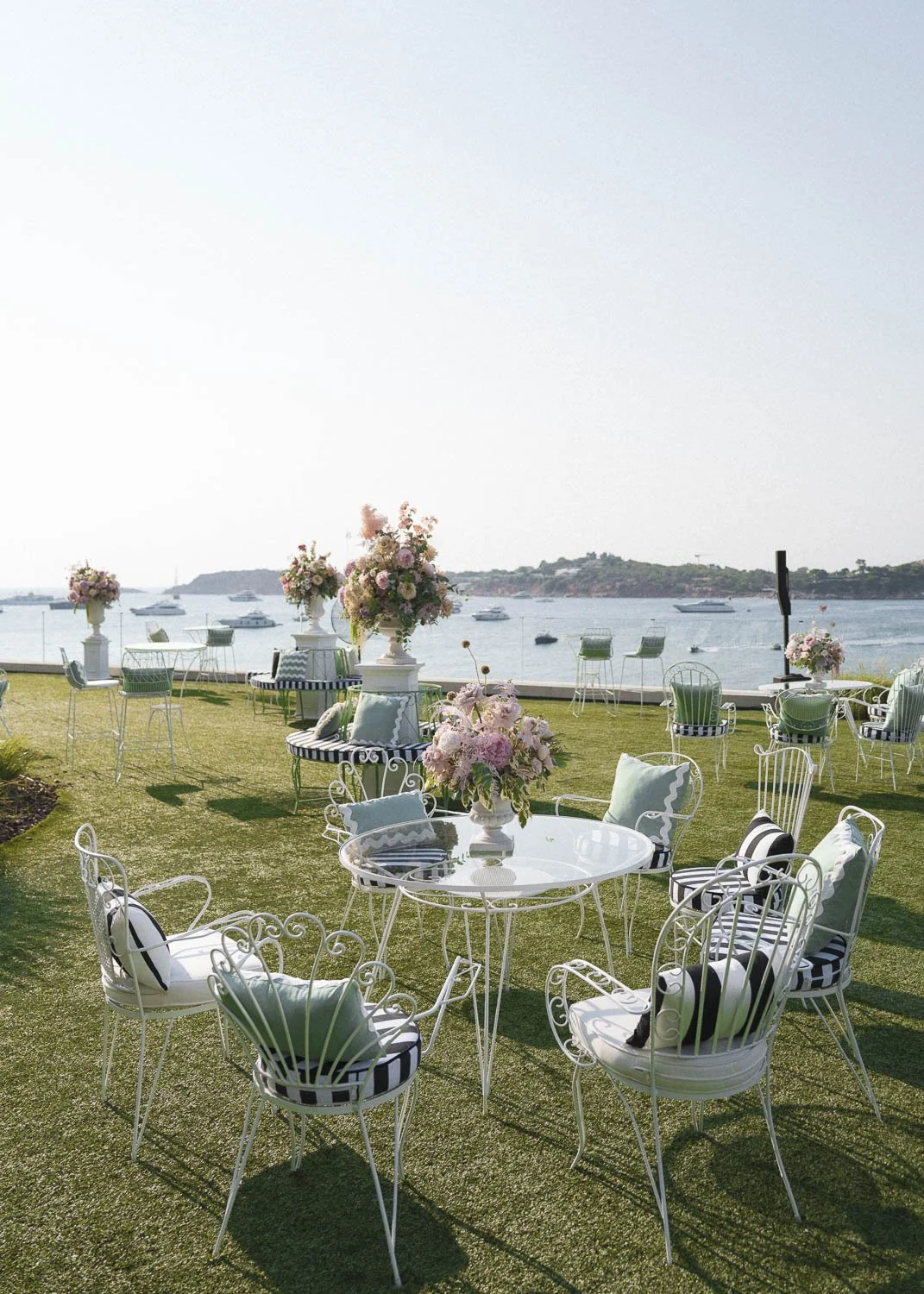 Outdoor event setup with white wrought-iron tables and chairs on a grassy area near water, decorated with pink and white floral centerpieces, overlooking boats and a distant shoreline.