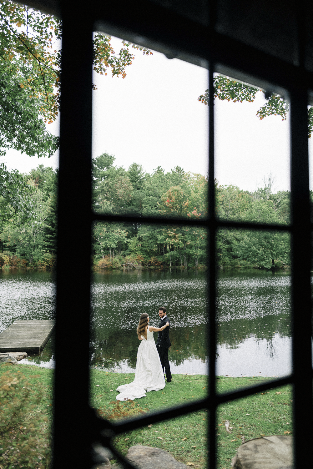 A bride and groom are dancing by a lake on a grassy area, seen through a window with multiple panes. The scene is surrounded by trees with green and some autumn-colored leaves.