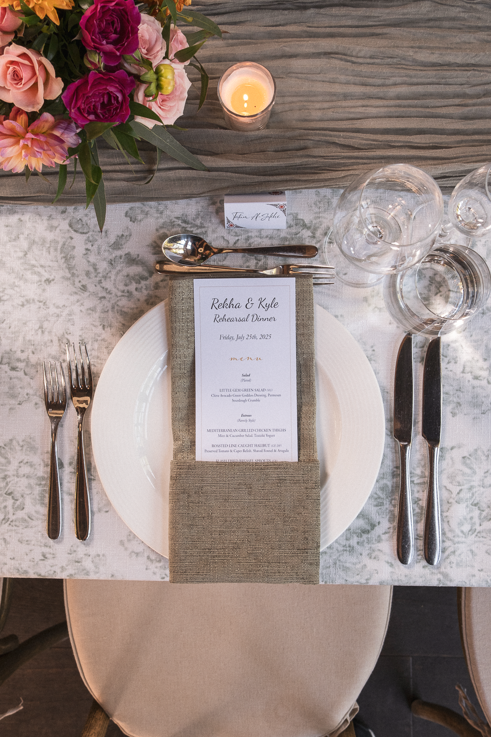 A formal dinner table setup with a floral centerpiece, a lit candle, a dinner plate with a menu card on a napkin, and glassware. The table is decorated with neutral tones and elegant silverware.