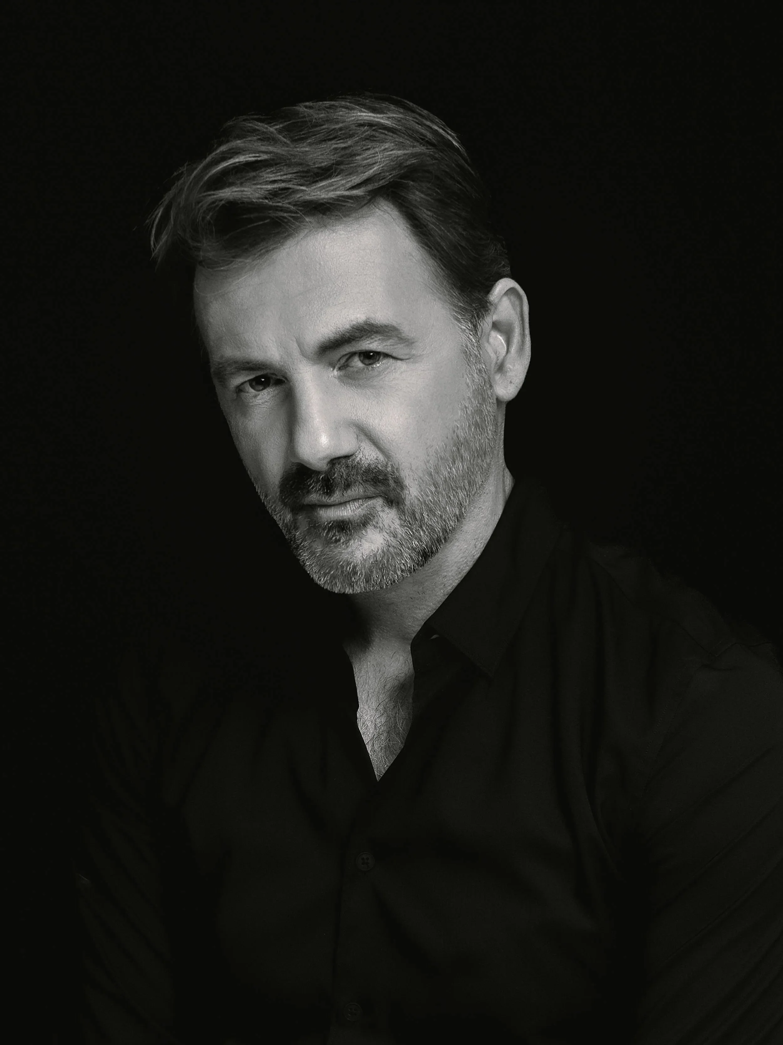 A black and white portrait of a man with styled hair, a beard, and wearing a dark shirt, looking directly at the camera against a dark background.