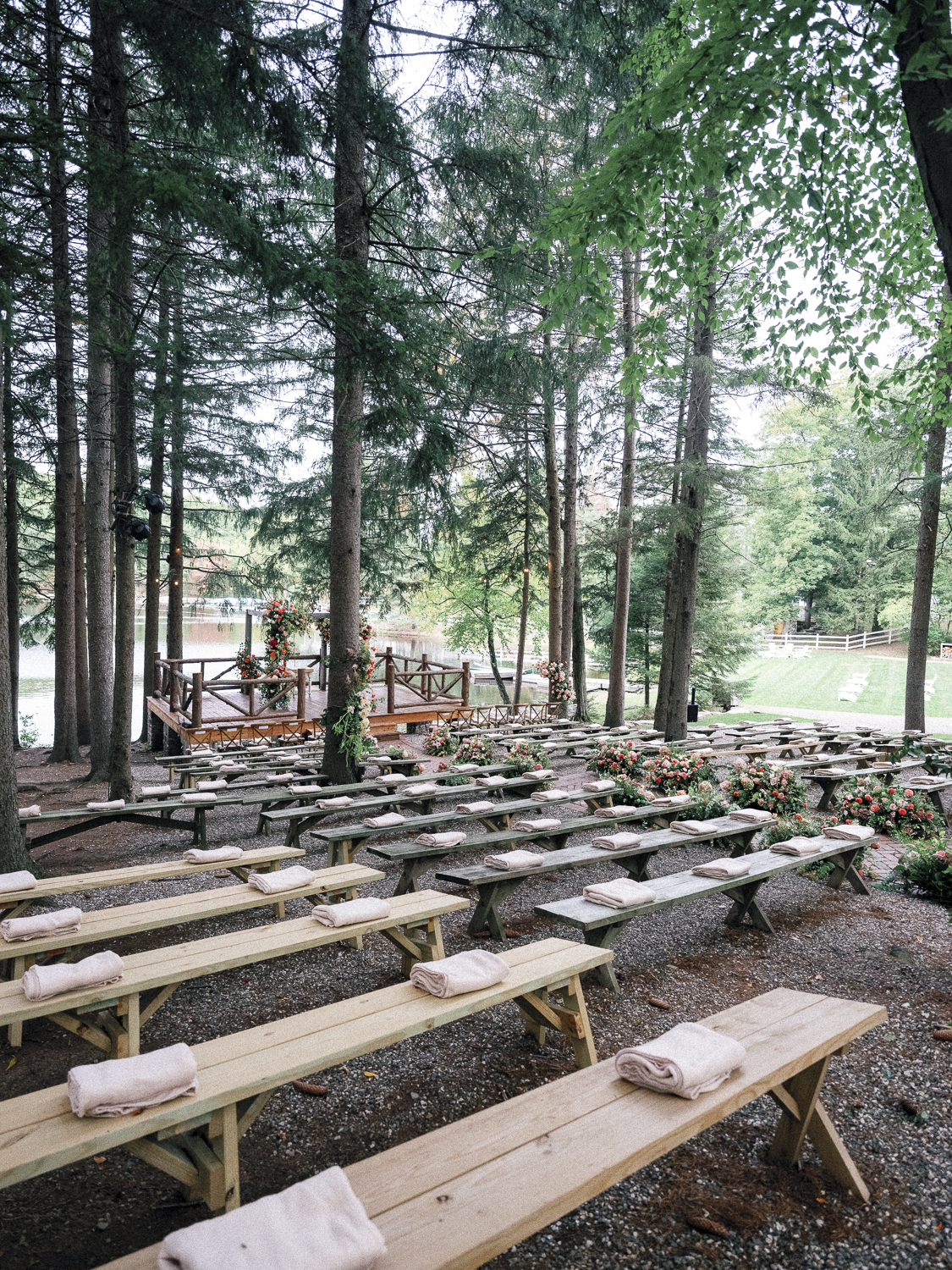 Outdoor wedding ceremony setup with wooden benches, folded towels, floral arrangements, and a small wooden platform surrounded by tall trees near a lake.
