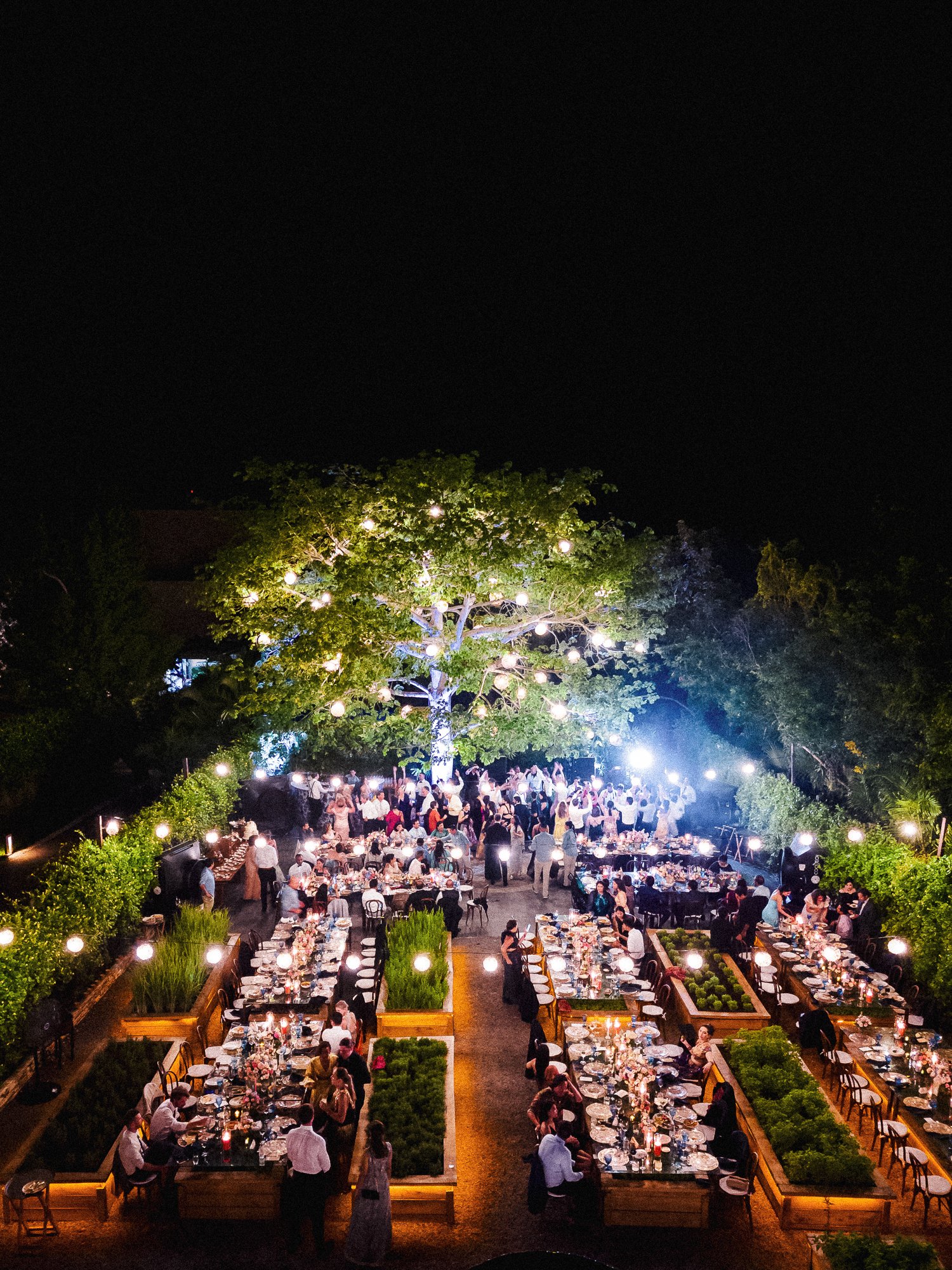 Outdoor nighttime event with numerous tables lit by candles, with guests mingling and dining under a lit tree decorated with hanging lanterns.