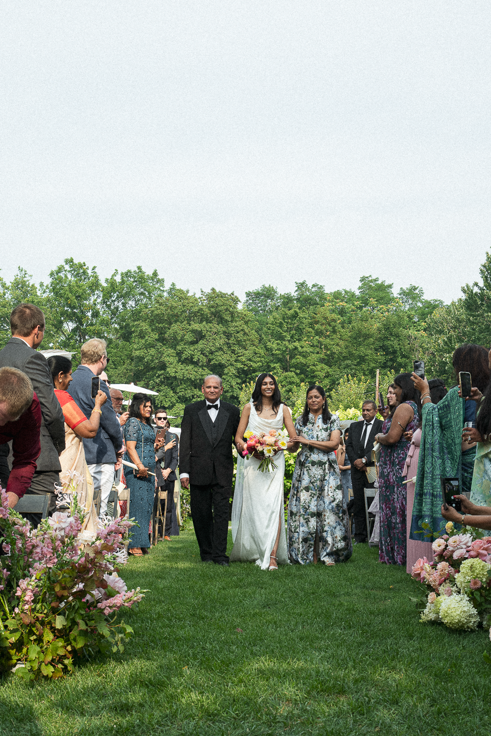 Bride walking down the aisle with her father at an outdoor wedding ceremony, surrounded by guests taking photos.