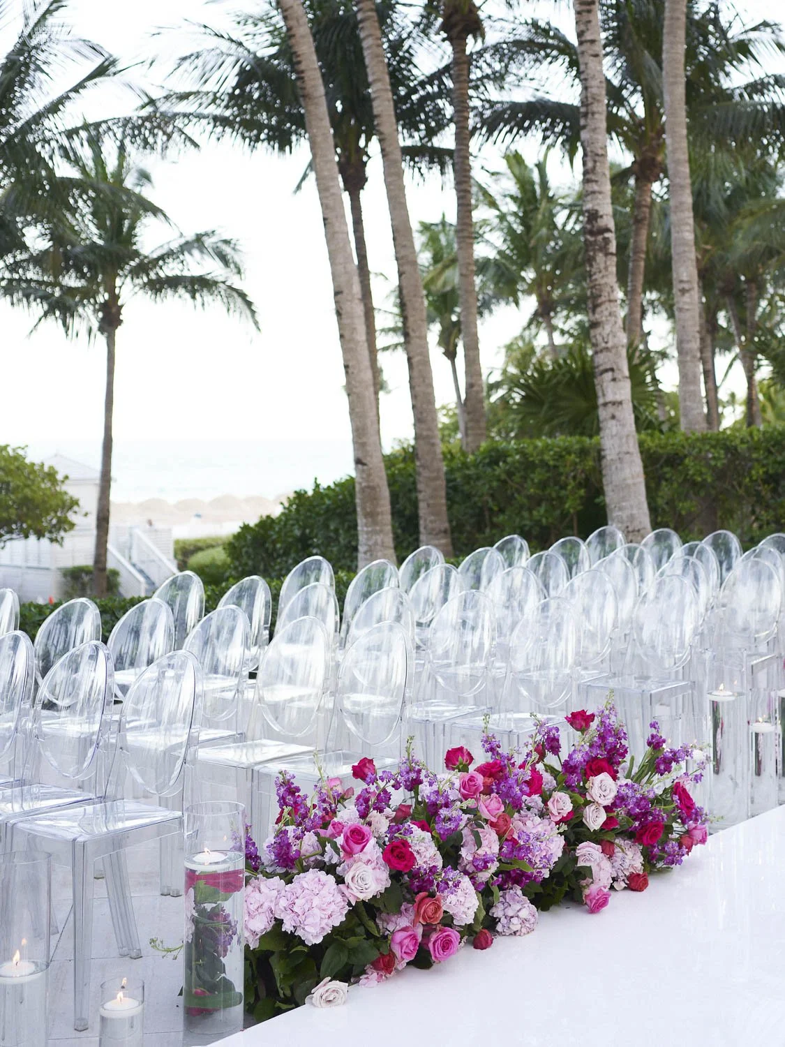 Outdoor wedding setup with clear acrylic chairs arranged in rows, decorated with a long floral arrangement of pink and purple flowers, surrounded by palm trees and greenery.