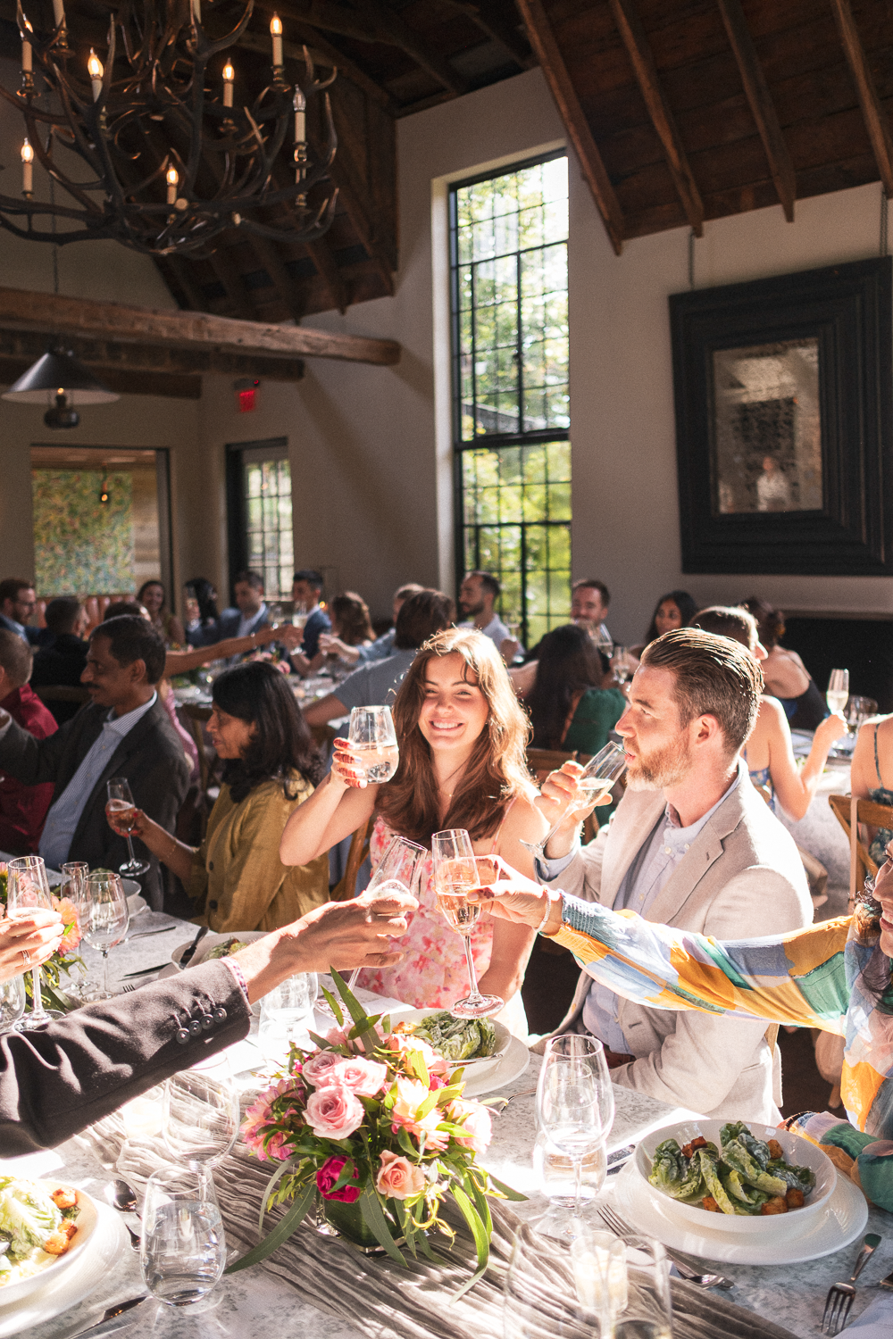 People celebrating at a long table with food and drinks in a rustic room with wooden beams and large windows, cheerful gathering with toasting