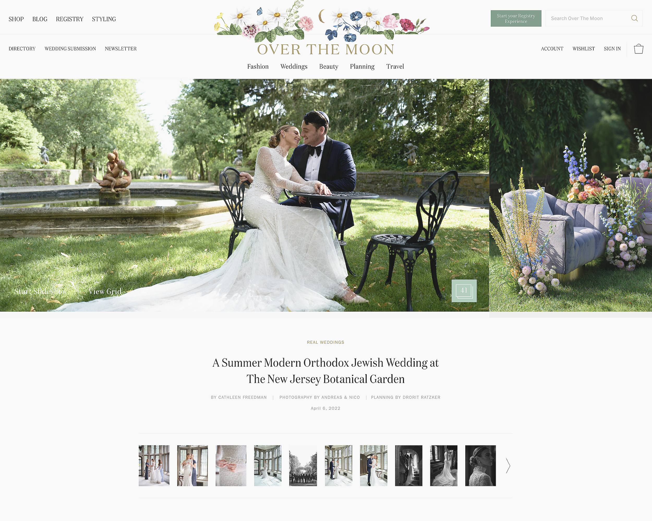 A couple in wedding attire seated at a black wrought-iron table outdoors in a lush garden with a fountain and statue in the background. The bride is in a white lace wedding dress, and the groom is in a navy tuxedo. A gray tufted sofa decorated with a