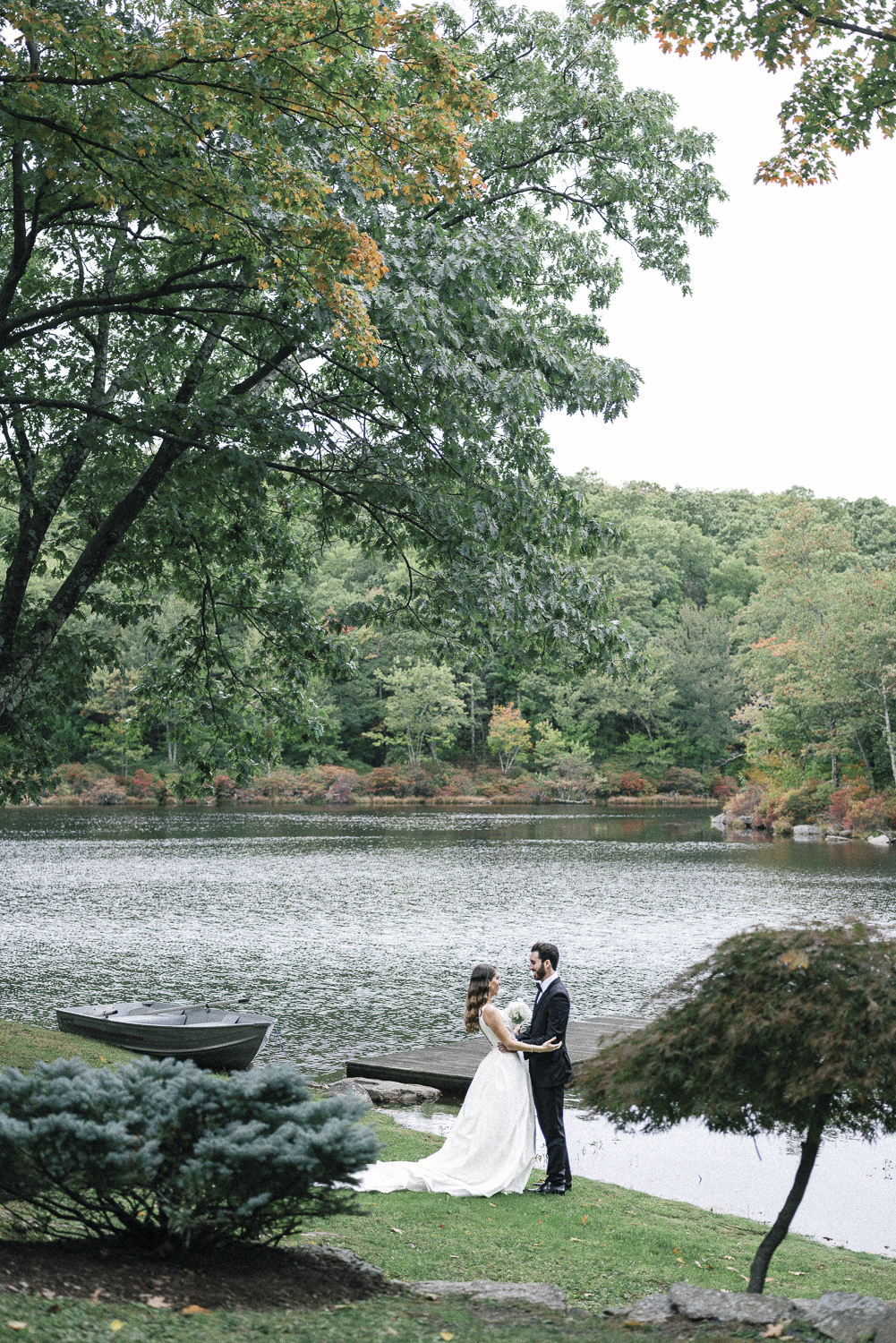 A bride and groom stand by a lake surrounded by trees, with the bride in a white wedding gown and the groom in a dark suit, holding hands and looking into each other's eyes.