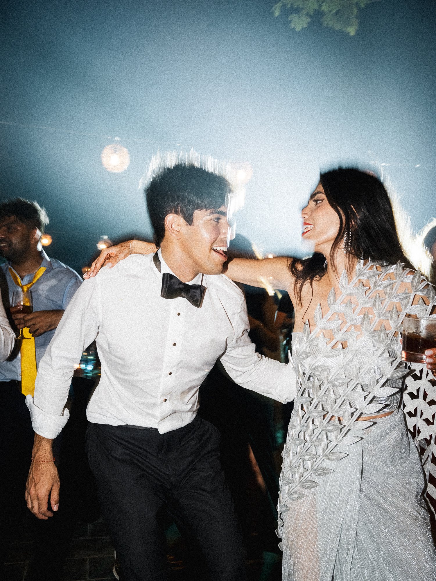 A man and woman dance closely at a celebration, smiling at each other, with the man in a tuxedo and the woman in a silver dress.