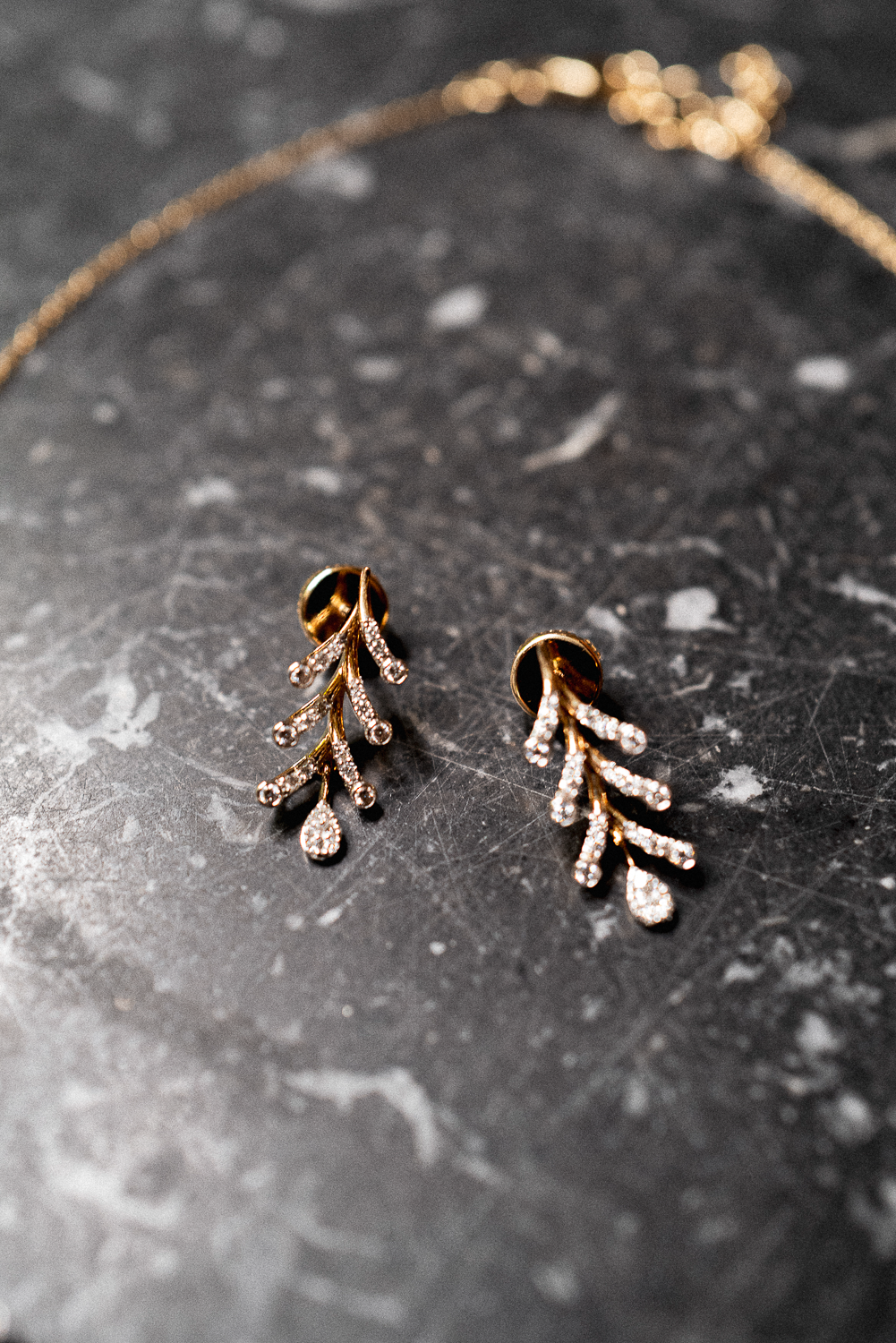 Gold leaf-shaped earrings with embedded diamonds resting on a dark, speckled surface.