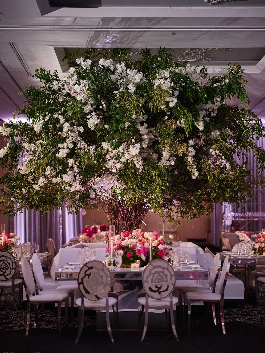 Large indoor event space decorated with a giant flowering tree centerpiece surrounded by elegant tables with pink and white floral centerpieces.