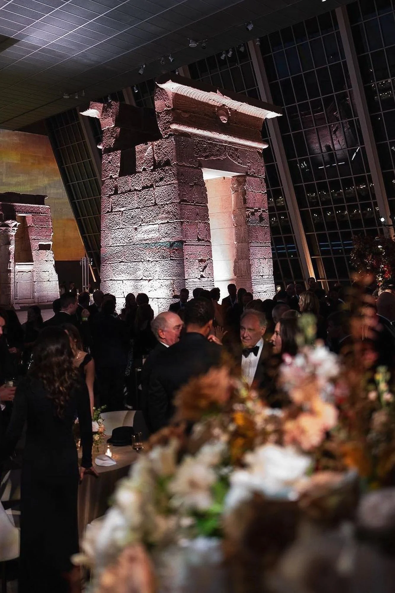 A large indoor event with many people dressed in formal attire, including tuxedos and evening gowns. The setting features ancient stone columns illuminated by soft lighting, with floor-to-ceiling windows and a city skyline visible in the background. 