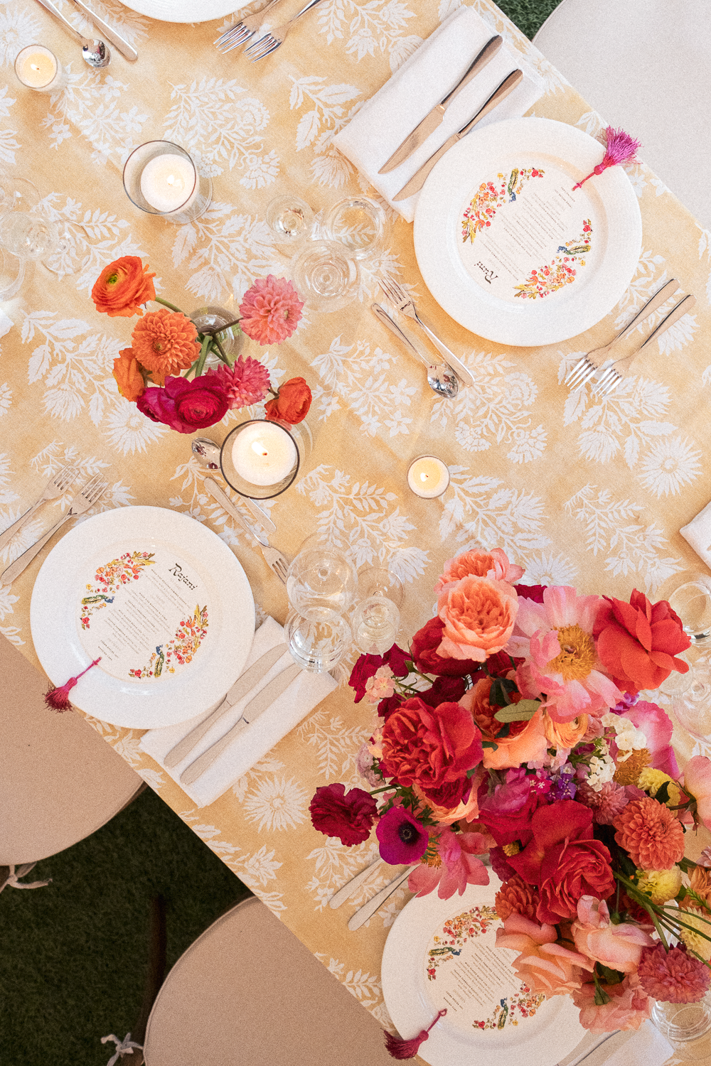 A table setting with floral arrangements, candles, and place settings with printed menus on elegant white plates, arranged on a cream-colored tablecloth with a leafy pattern.