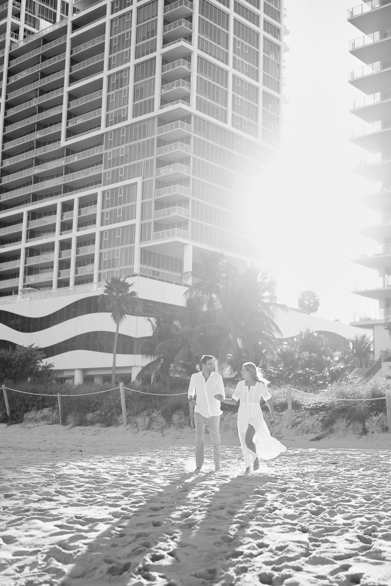 A couple walking on a sandy beach, holding hands and smiling at each other, with tall modern apartment buildings in the background and palm trees nearby.
