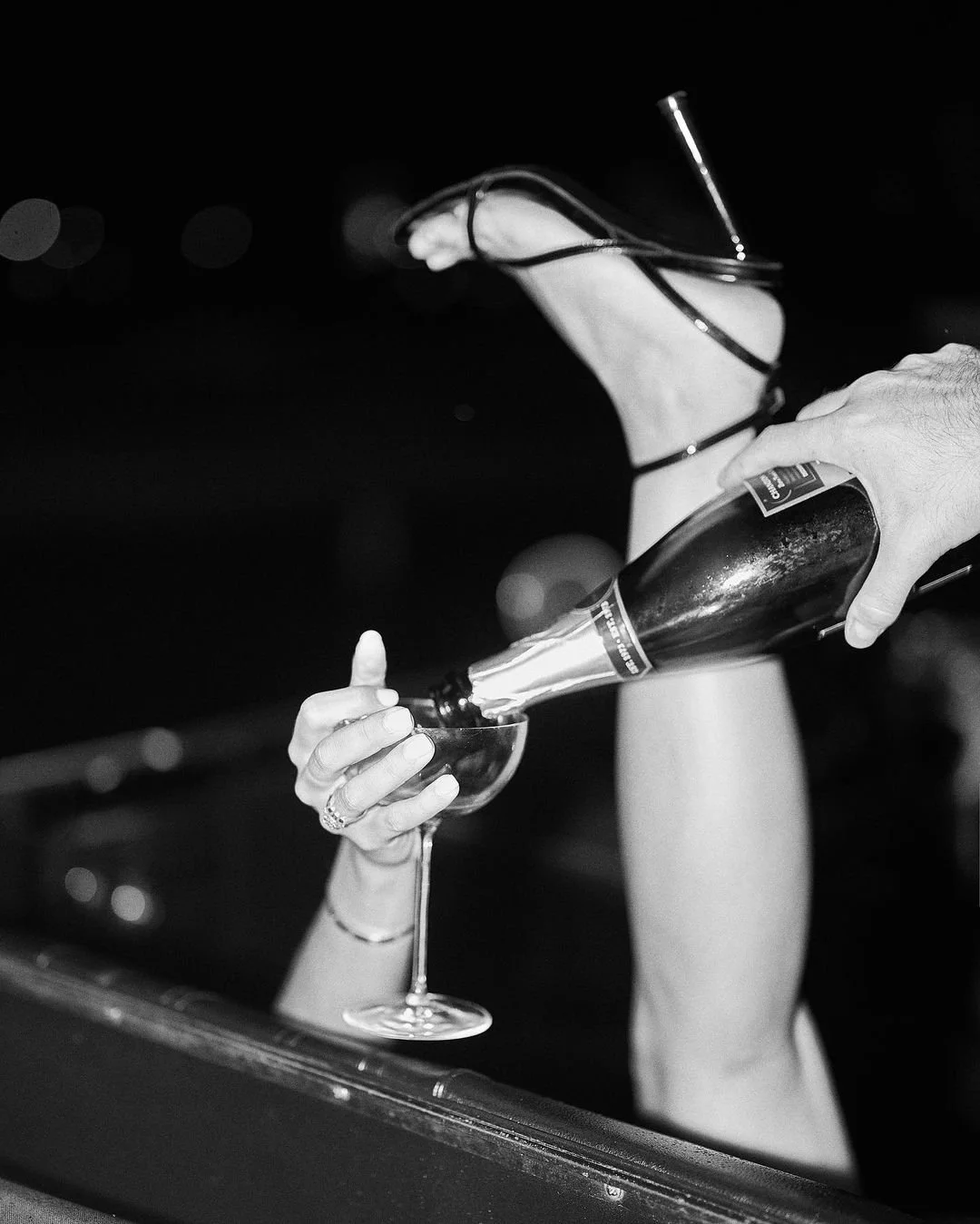 A woman with manicured nails is holding a champagne glass, while a hand is pouring champagne into it from a bottle.