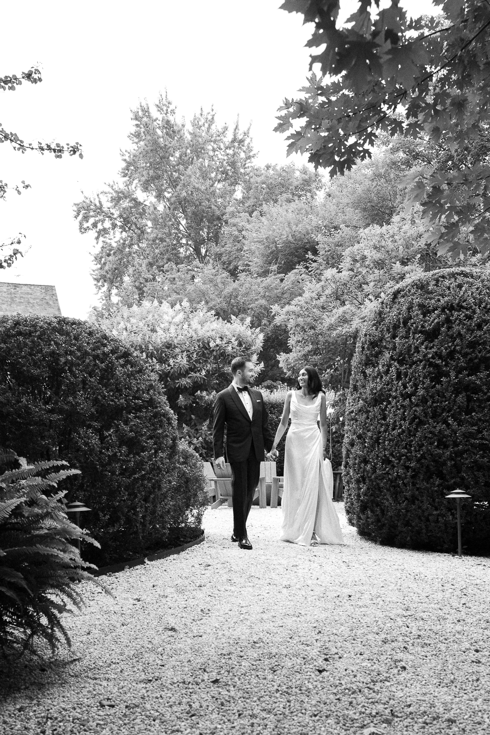 A black and white photo of a couple walking hand in hand through a garden, the man in a tuxedo and the woman in a long gown, surrounded by tall bushes and trees.
