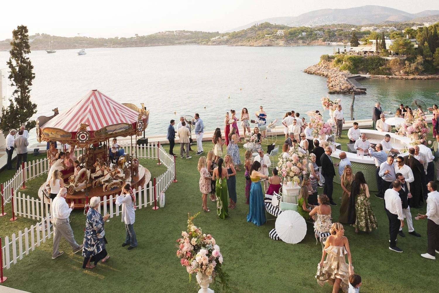 A lively outdoor celebration by the water with a carousel, pink floral decorations, and guests dressed in colorful attire enjoying the event at sunset.