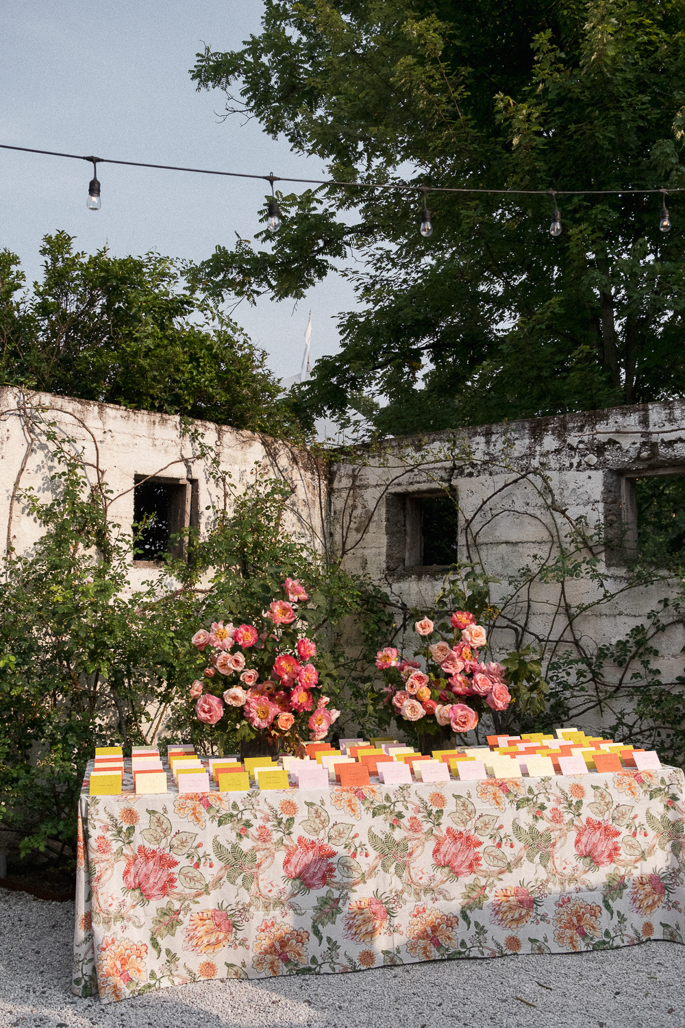 Outdoor table with floral tablecloth, floral arrangements, and colorful cards, set against a weathered white brick wall with vines, surrounded by trees, with string lights overhead.