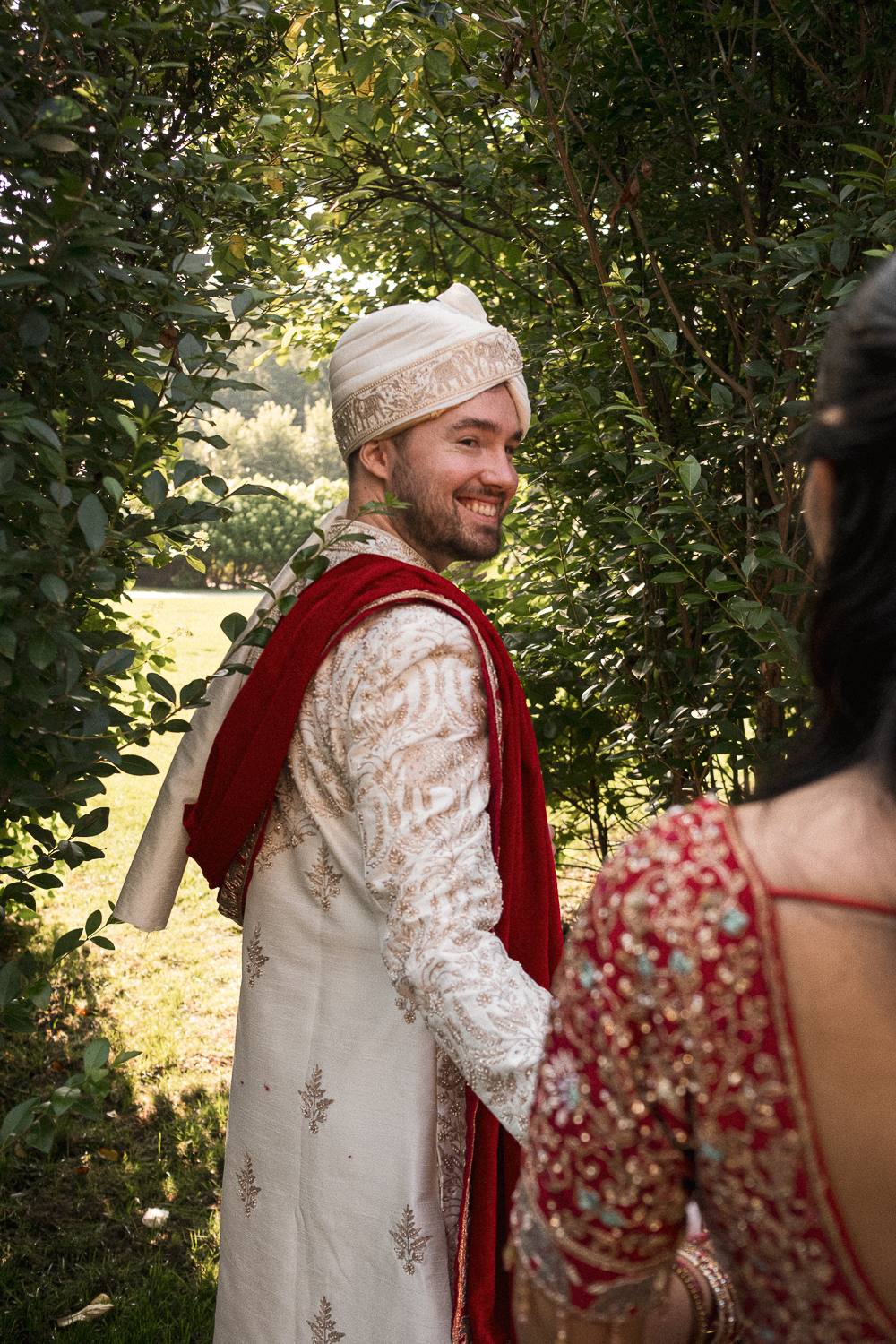 A man dressed in traditional Indian wedding attire, including a cream-colored sherwani with intricate gold embroidery, a matching turban, and a red dupatta, smiling as he looks back towards a woman in a red and gold embroidered outfit in an outdoor s