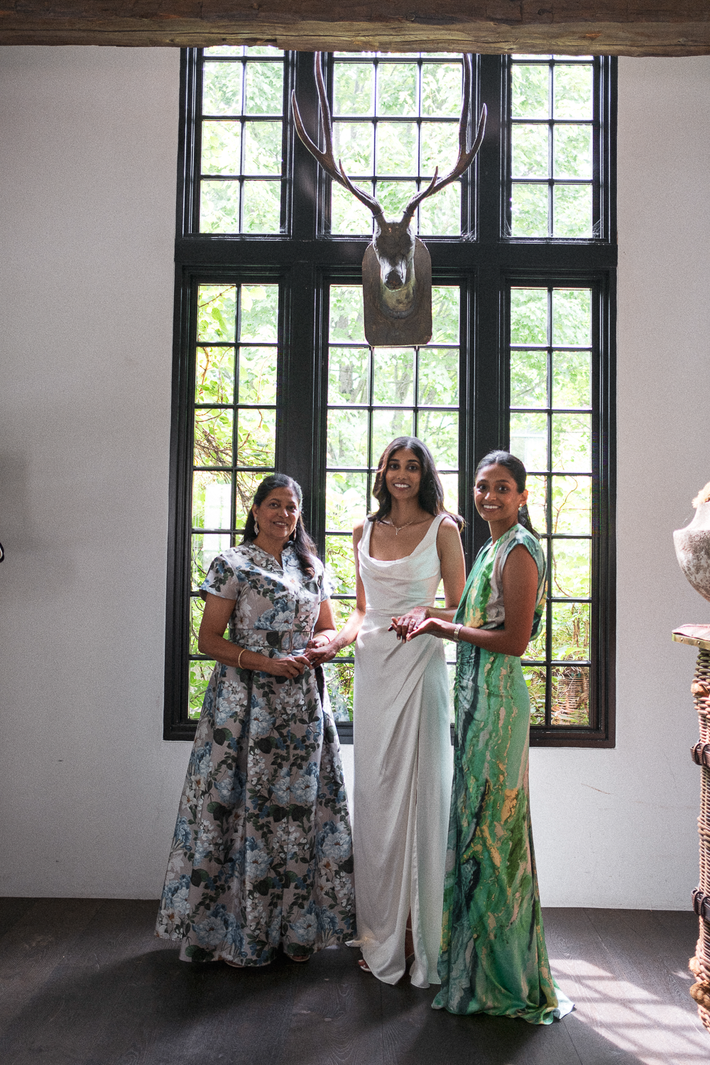 Three women standing inside in front of a large window with green trees outside, with a mounted deer head on the window frame.