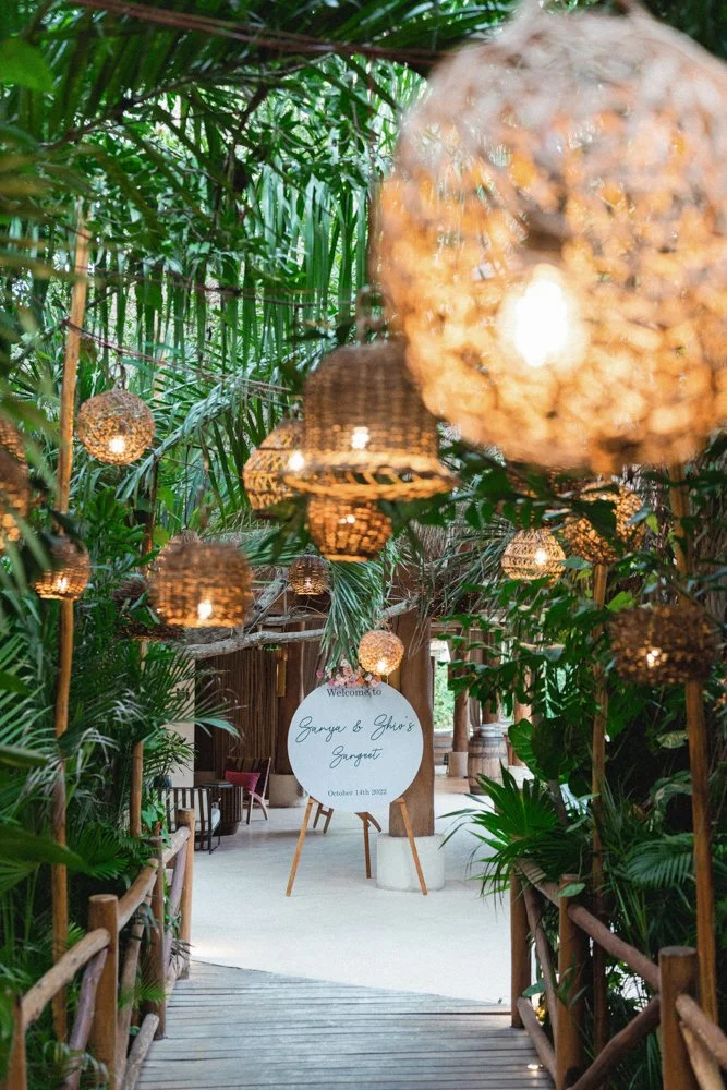 Pathway with hanging woven lamps and lush tropical greenery leading to a sign that reads, 'Welcome to Ganya & Giny's Garden, October 14th, 2022'.