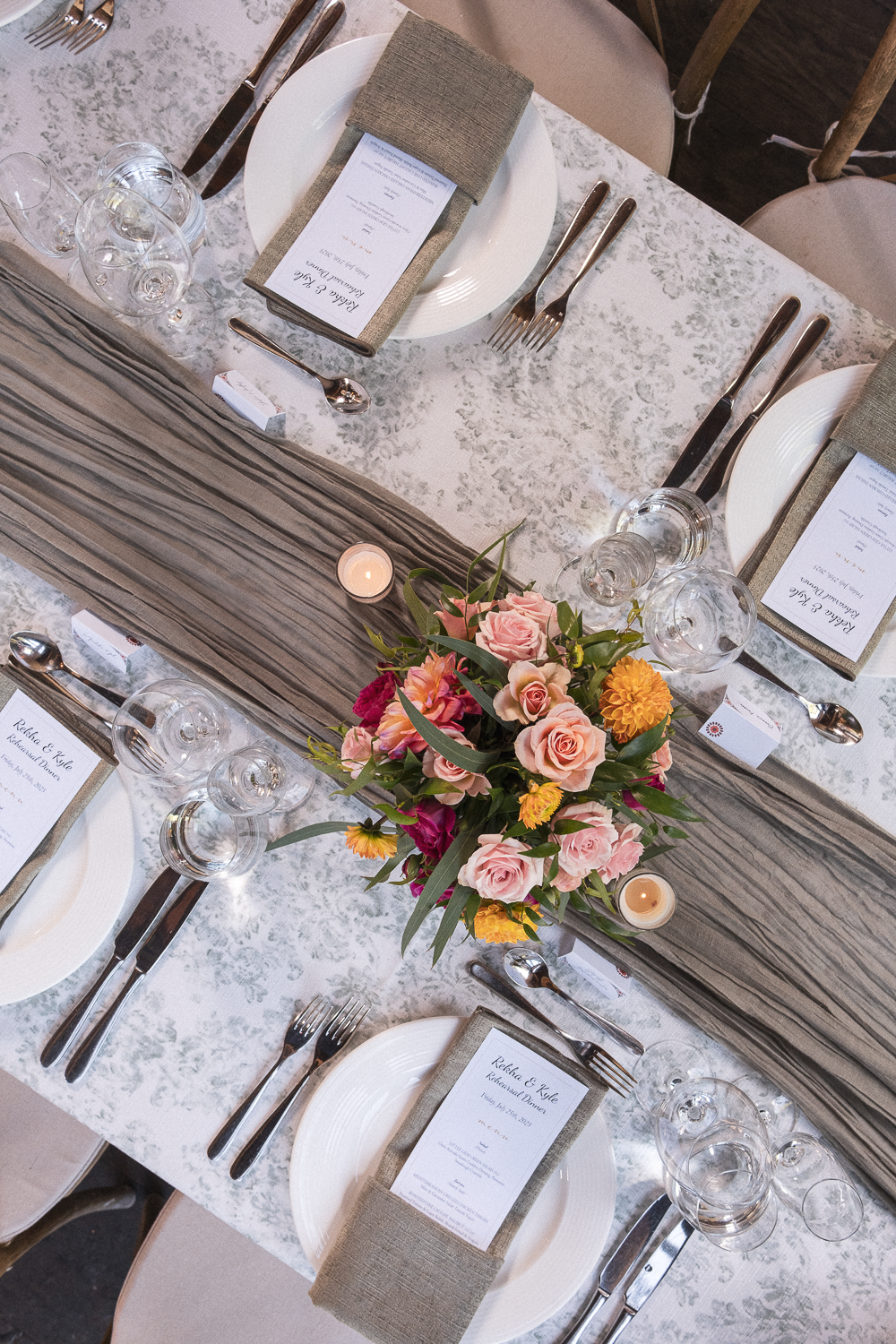 A formal dining table setup with white plates, silverware, wine glasses, and beige napkins. There's a gray table runner and a pink, peach, and yellow floral centerpiece with candles around it. Guests' name cards are on the plates.