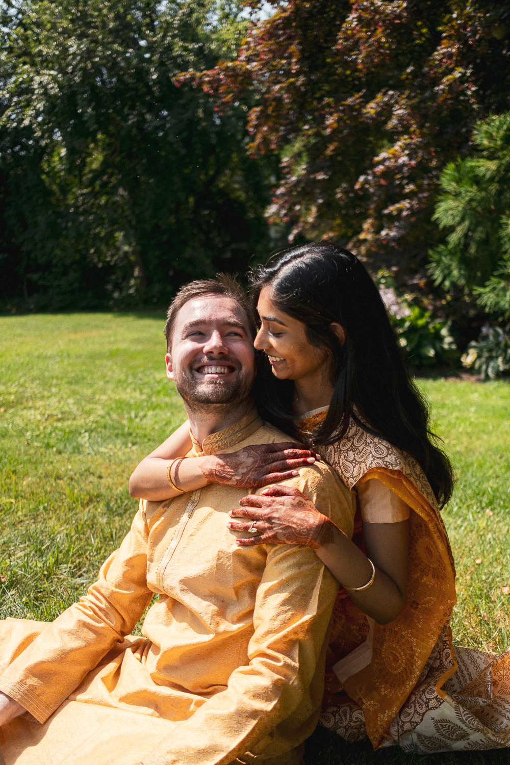 A happy couple sitting on grass in traditional Indian attire, embracing and smiling outdoors with trees in the background.