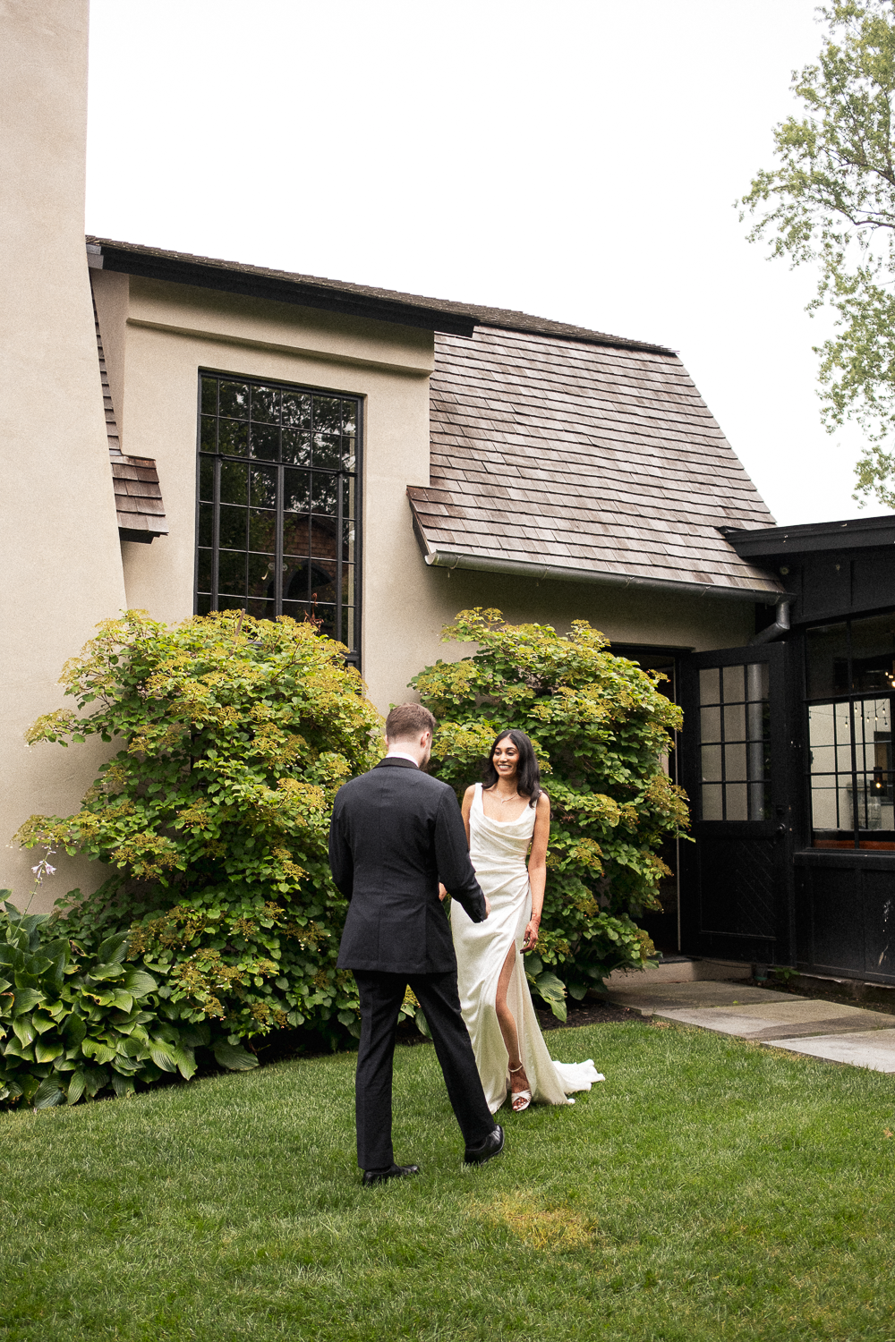 A couple dressed in wedding attire standing on a green lawn outside a modern house with large windows and lush bushes in the background.