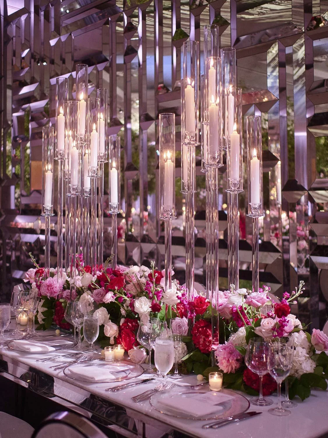 Elegant banquet table with tall glass candle holders, pink and red floral arrangements, and candles, set in a decorative venue with mirrored geometric walls.
