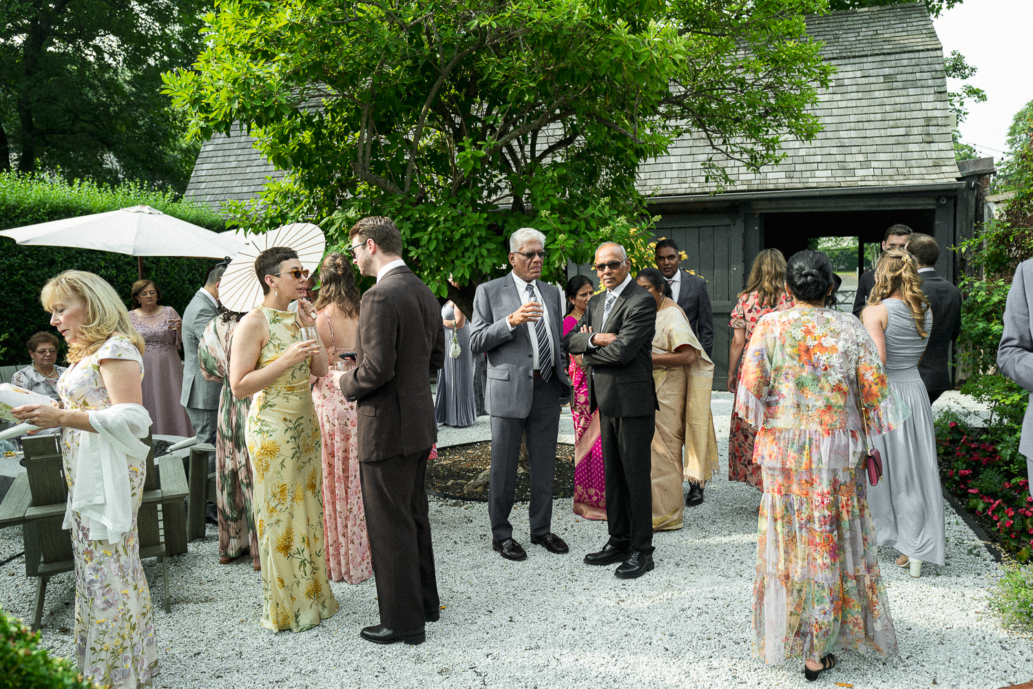 A group of people at an outdoor social gathering, dressed in formal and semi-formal attire, standing on gravel in front of a small black wooden building, with trees and greenery around, and a white umbrella providing shade.