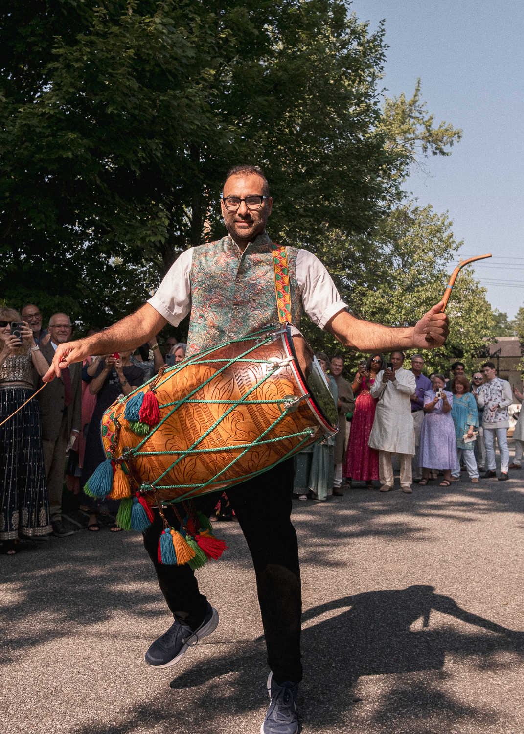 A man dancing with a dhol (drum) during a cultural celebration or parade, surrounded by a crowd of people taking photos and watching.