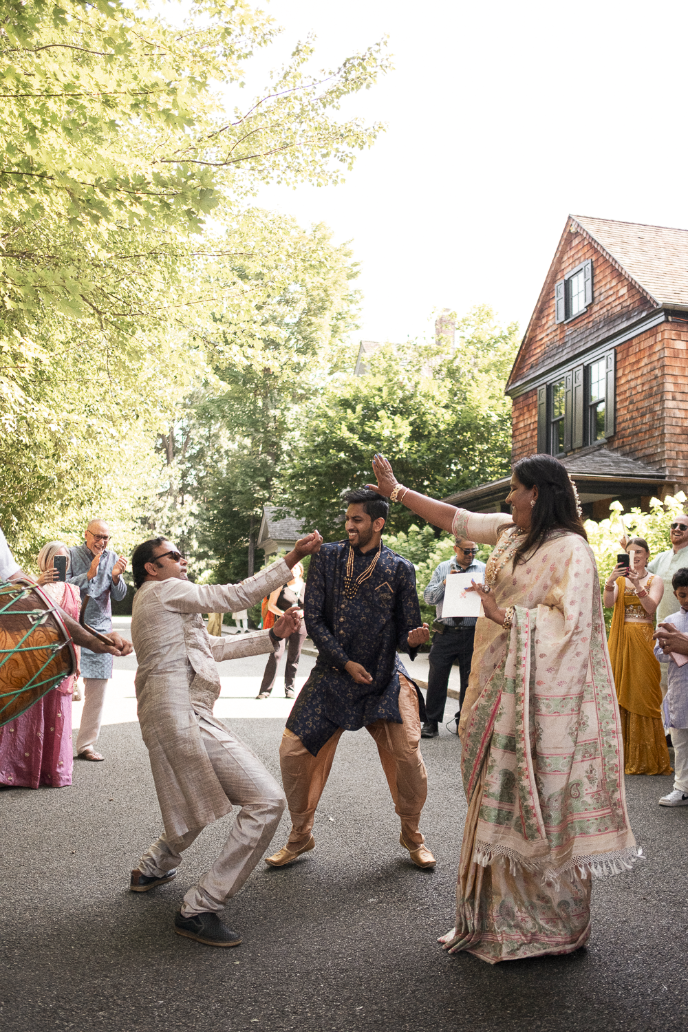 People dancing and celebrating outdoors in traditional Indian attire during a festive event, with a background of trees and a house.