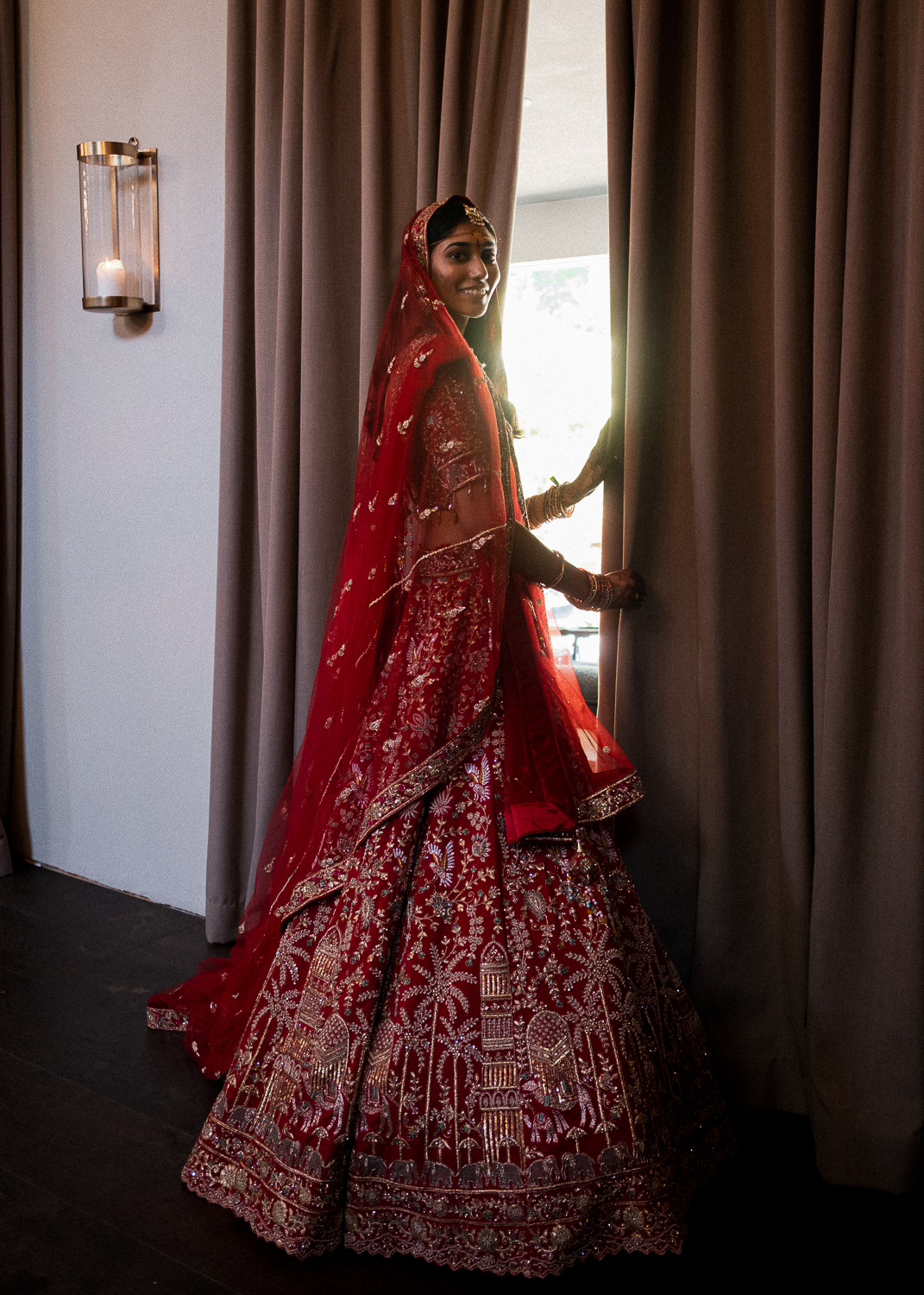 A woman in a traditional red Indian wedding dress, standing by a curtain, smiling and holding the curtains open.