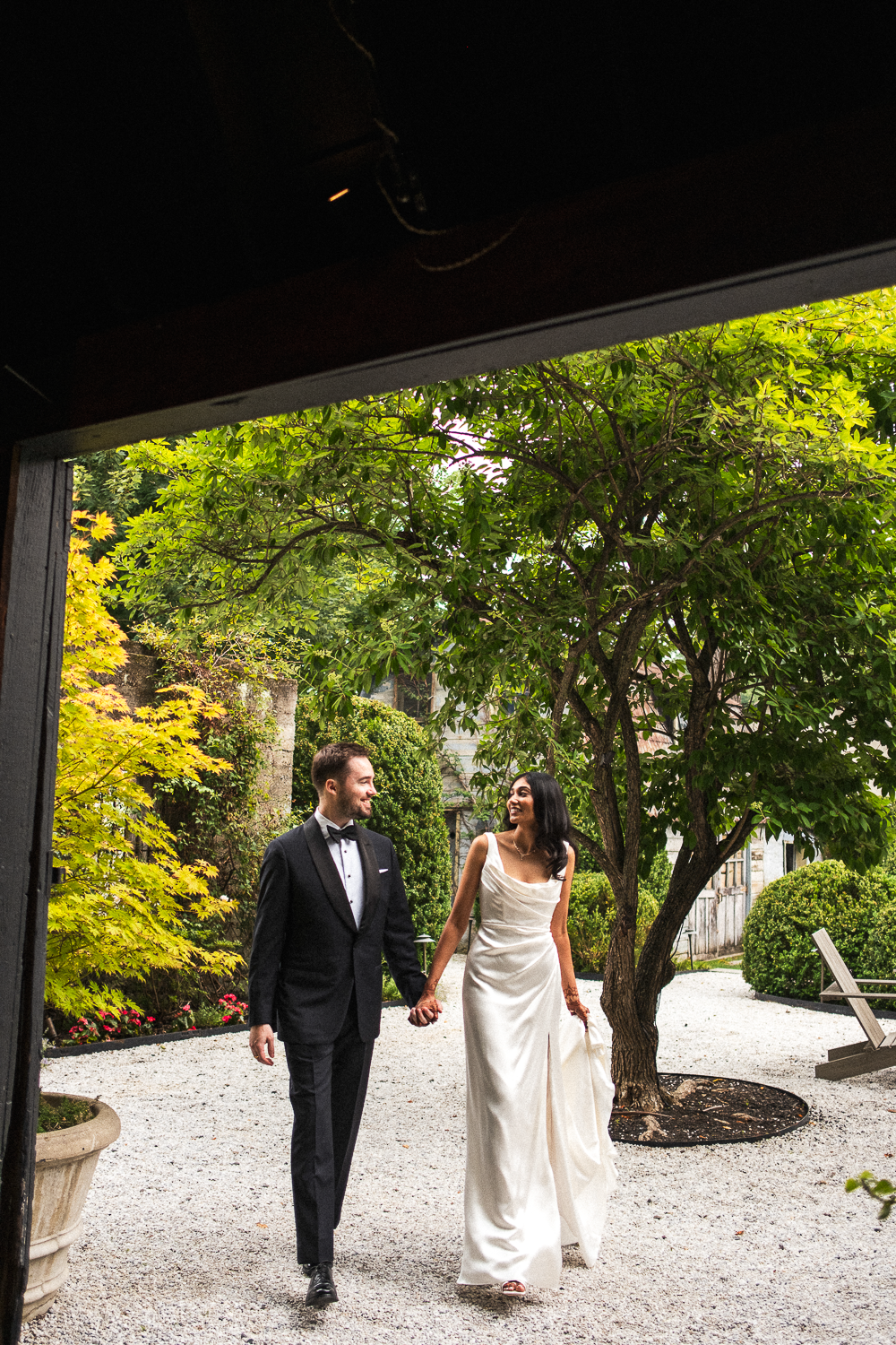 A bride and groom in wedding attire, holding hands and smiling at each other, walking outdoors in a lush garden with trees and shrubs.